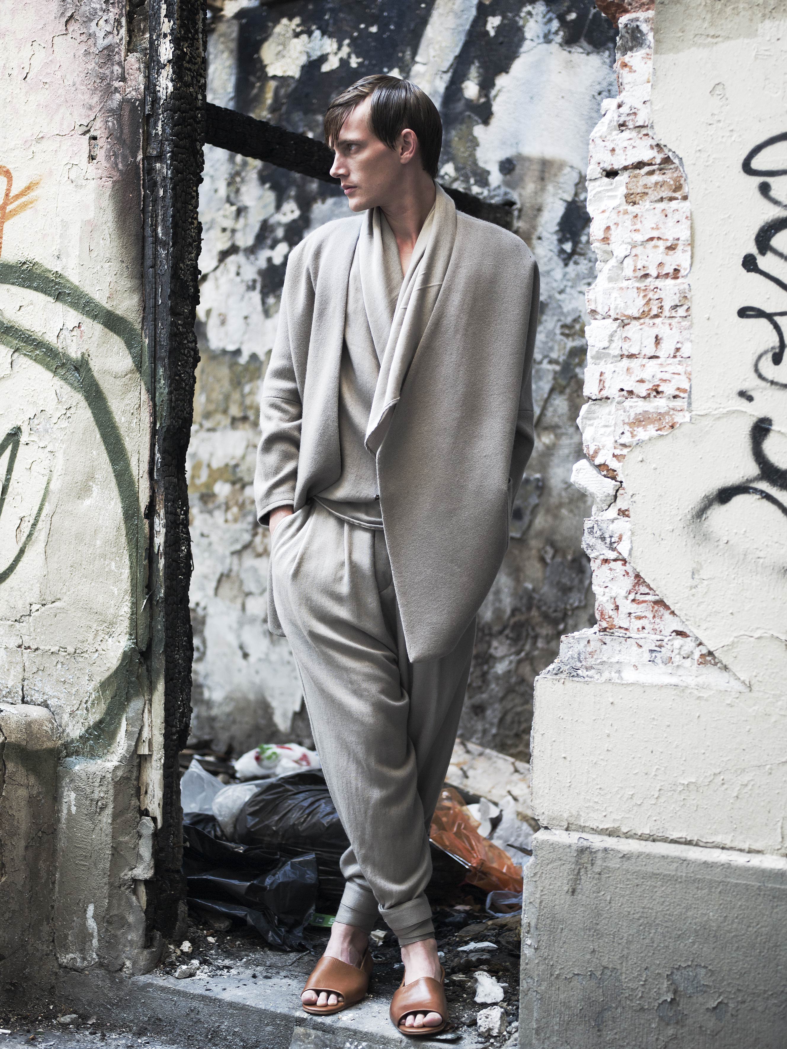 A person in a stylish, draped beige outfit stands against a weathered, graffiti-covered wall. Their hands are in the pockets of the loose-fitting ensemble, and they are looking to the side, wearing brown sandals. The surroundings are urban and rustic.