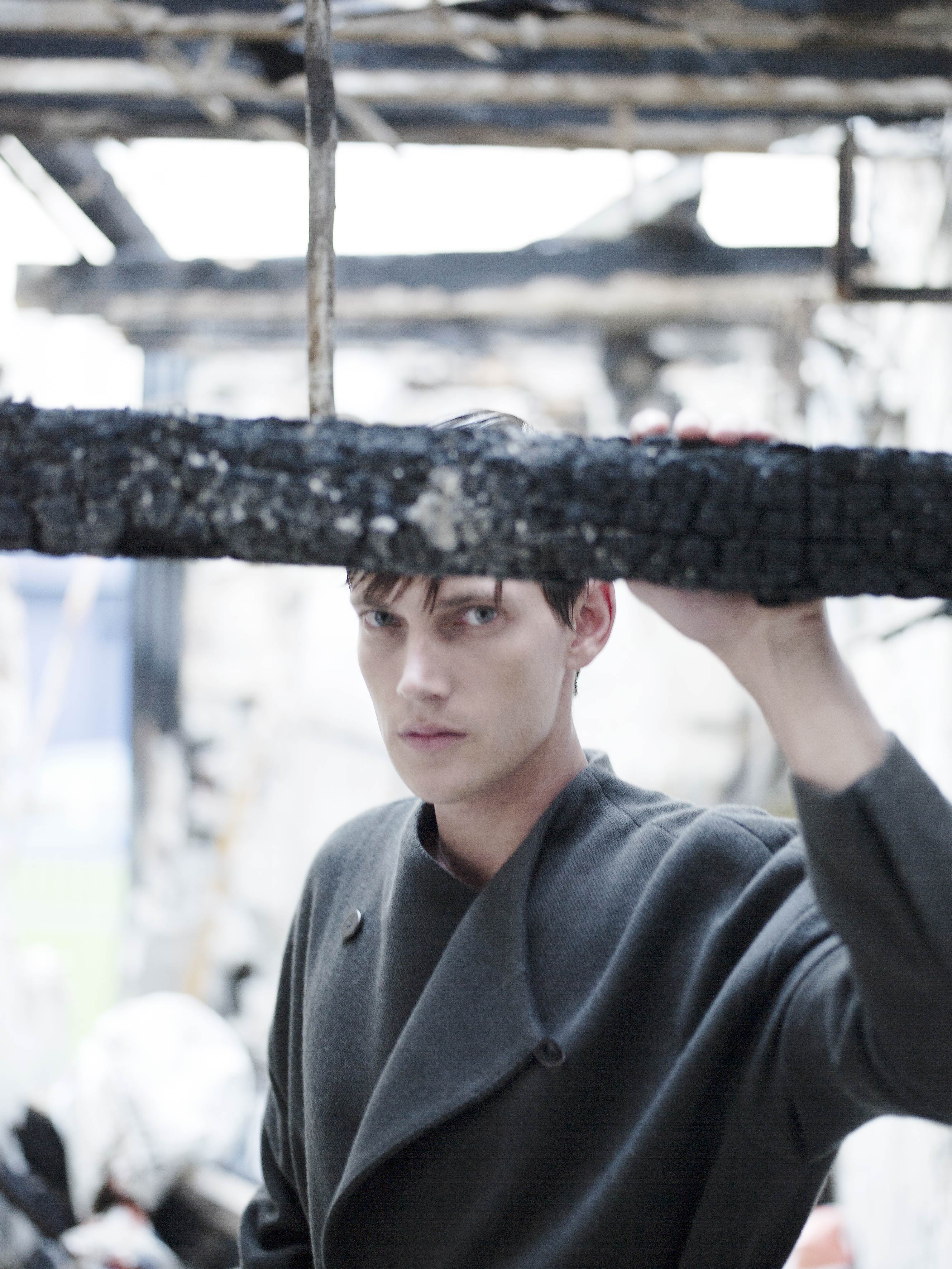 A person in a dark coat stands in a burned-out building, holding a piece of charred wood. The surroundings are damaged, with debris visible. The atmosphere is somber and reflective.