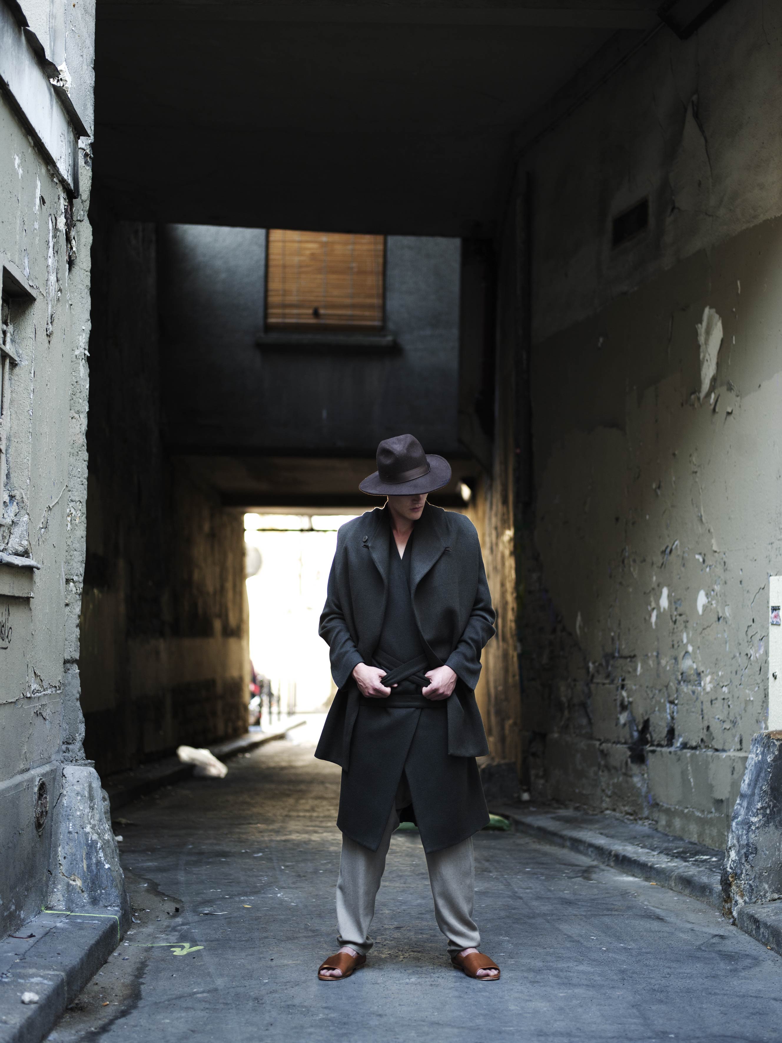 A person dressed in a dark coat, beige pants, and brown shoes stands in a dimly lit, narrow alley with peeling walls. They are wearing a wide-brimmed hat and looking down, with light coming from the alley entrance.