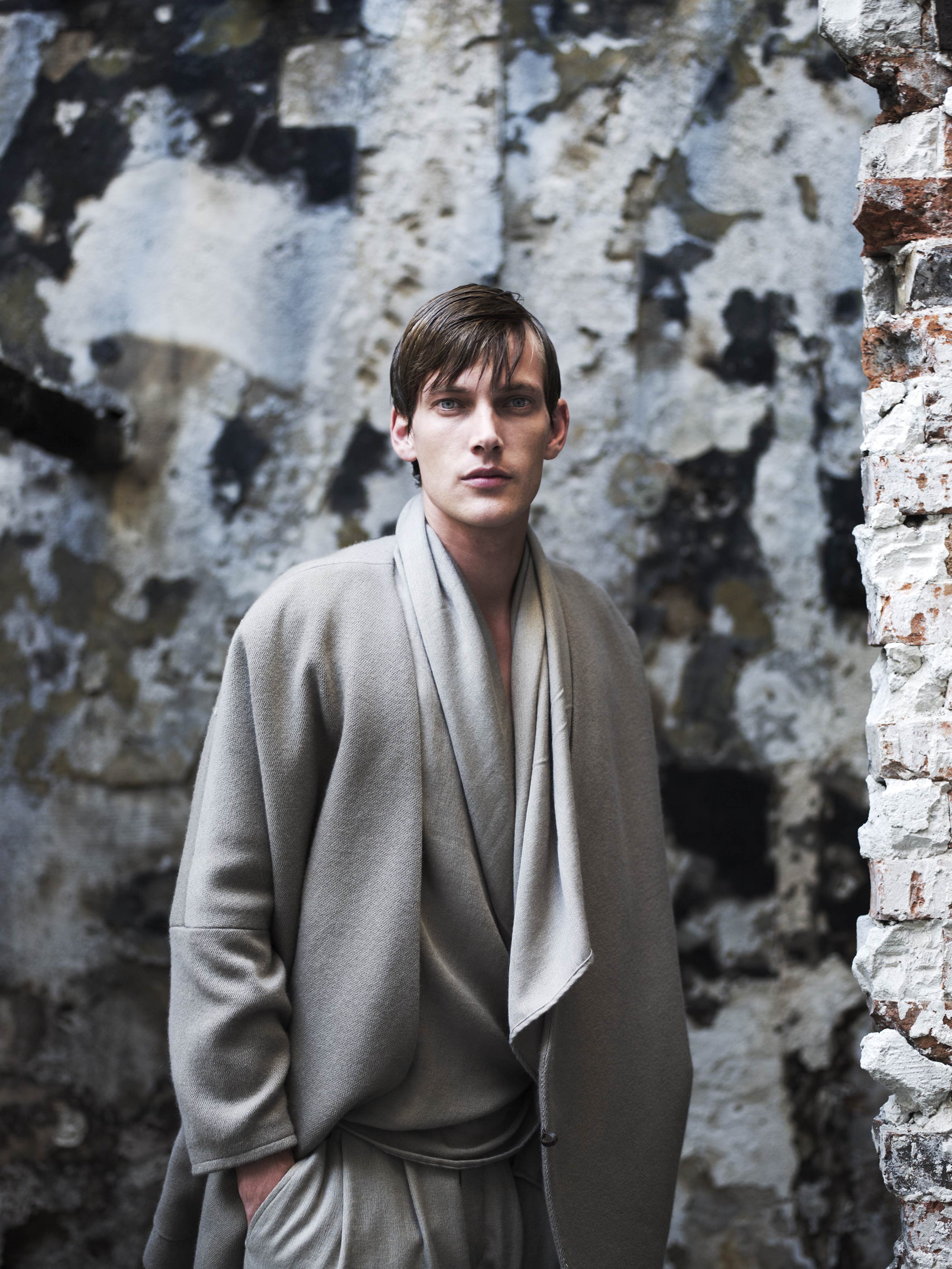 A person in a loose-fitting, earth-toned outfit stands in front of a weathered stone wall. The background features peeling paint and exposed brick, creating a rustic and textured setting.