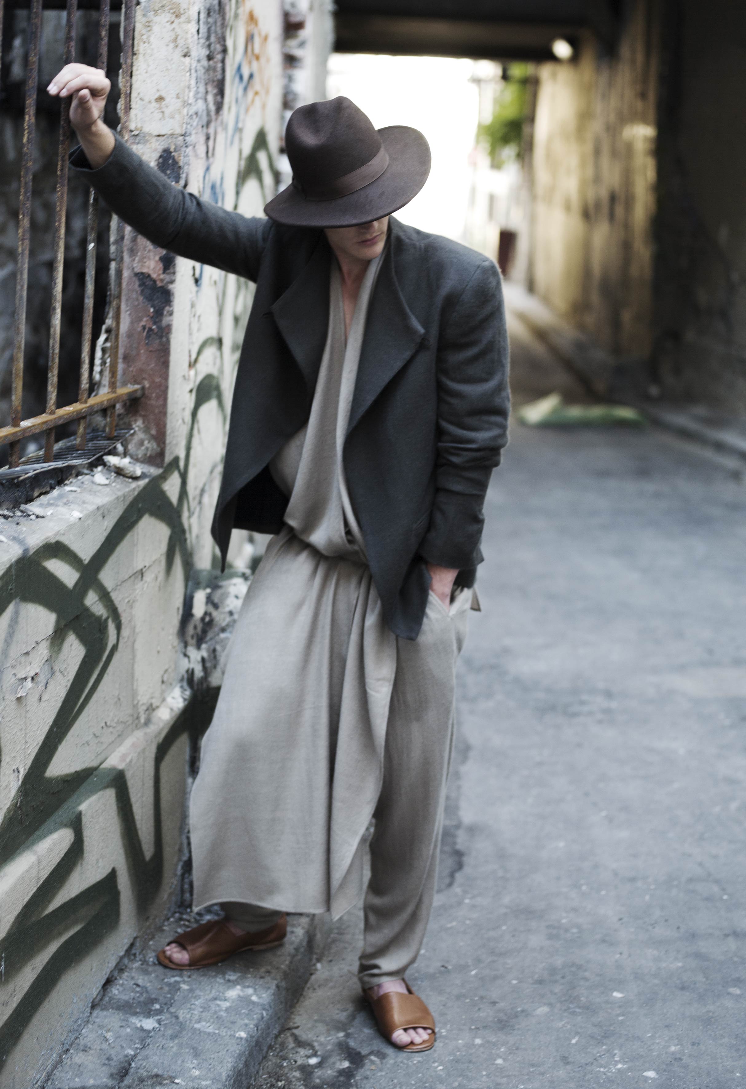 Person in a wide-brimmed hat and flowing beige outfit leans casually against a graffiti-covered wall in an urban alley. They wear a dark jacket and brown sandals, with a relaxed posture, looking down.