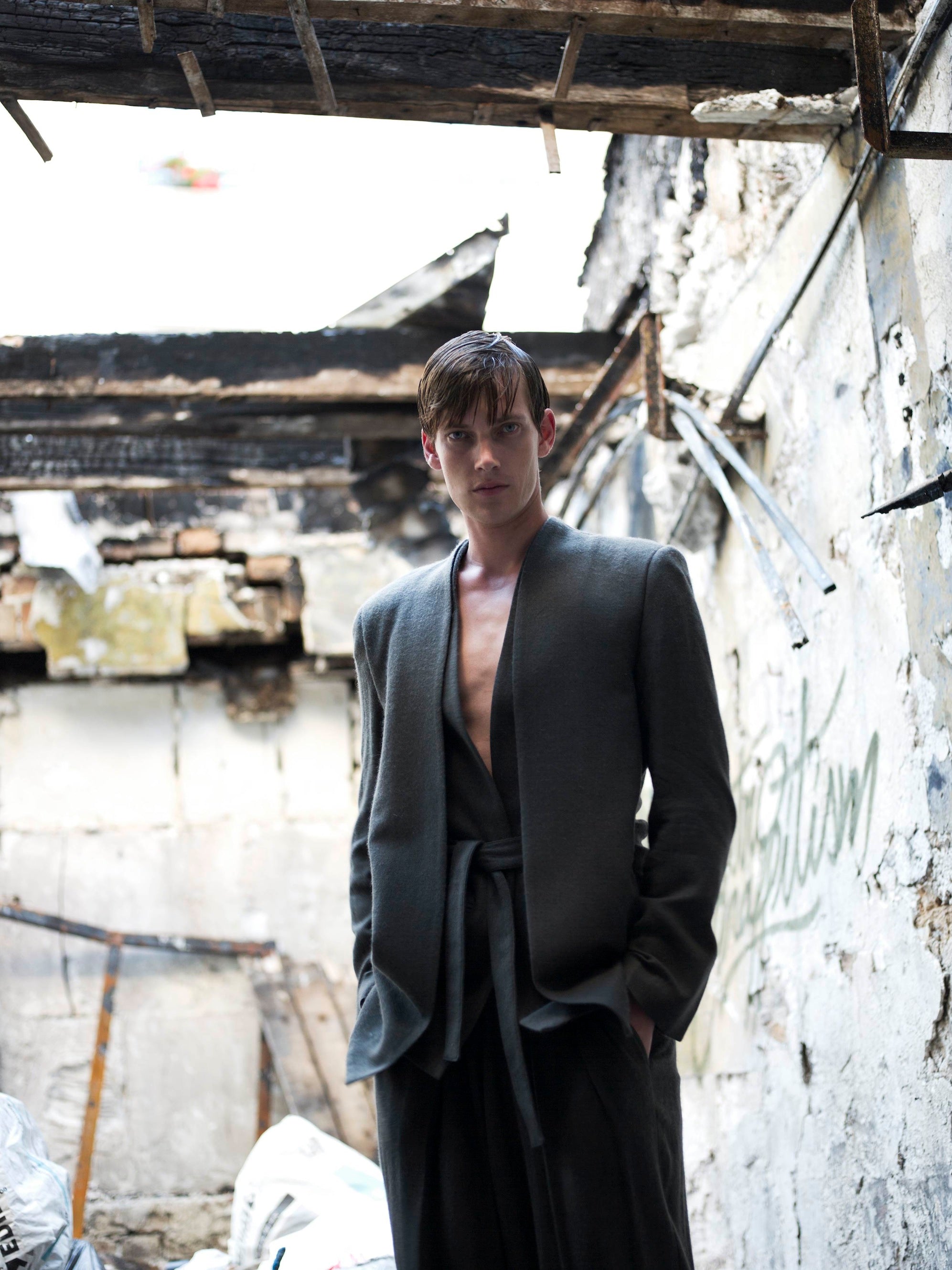 A person stands in a dilapidated building with exposed beams. They are wearing a dark, open jacket and relaxed trousers. The walls are crumbling, and debris is scattered on the ground. The sky is visible through the broken roof.