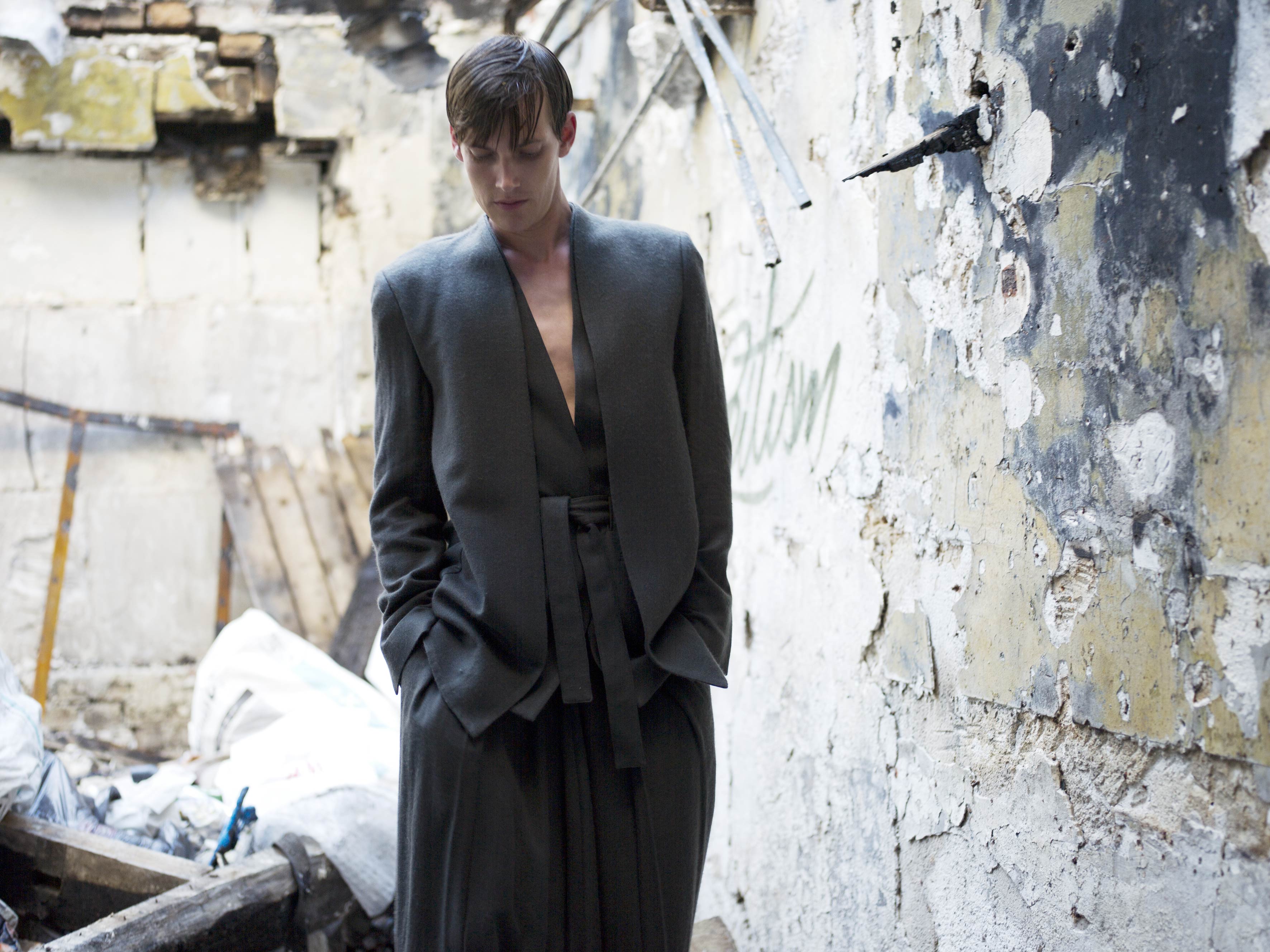 A person in a fashionable dark outfit stands with hands in pockets, looking down. They are in a dilapidated building with crumbling walls and scattered debris. The atmosphere is moody and contemplative.