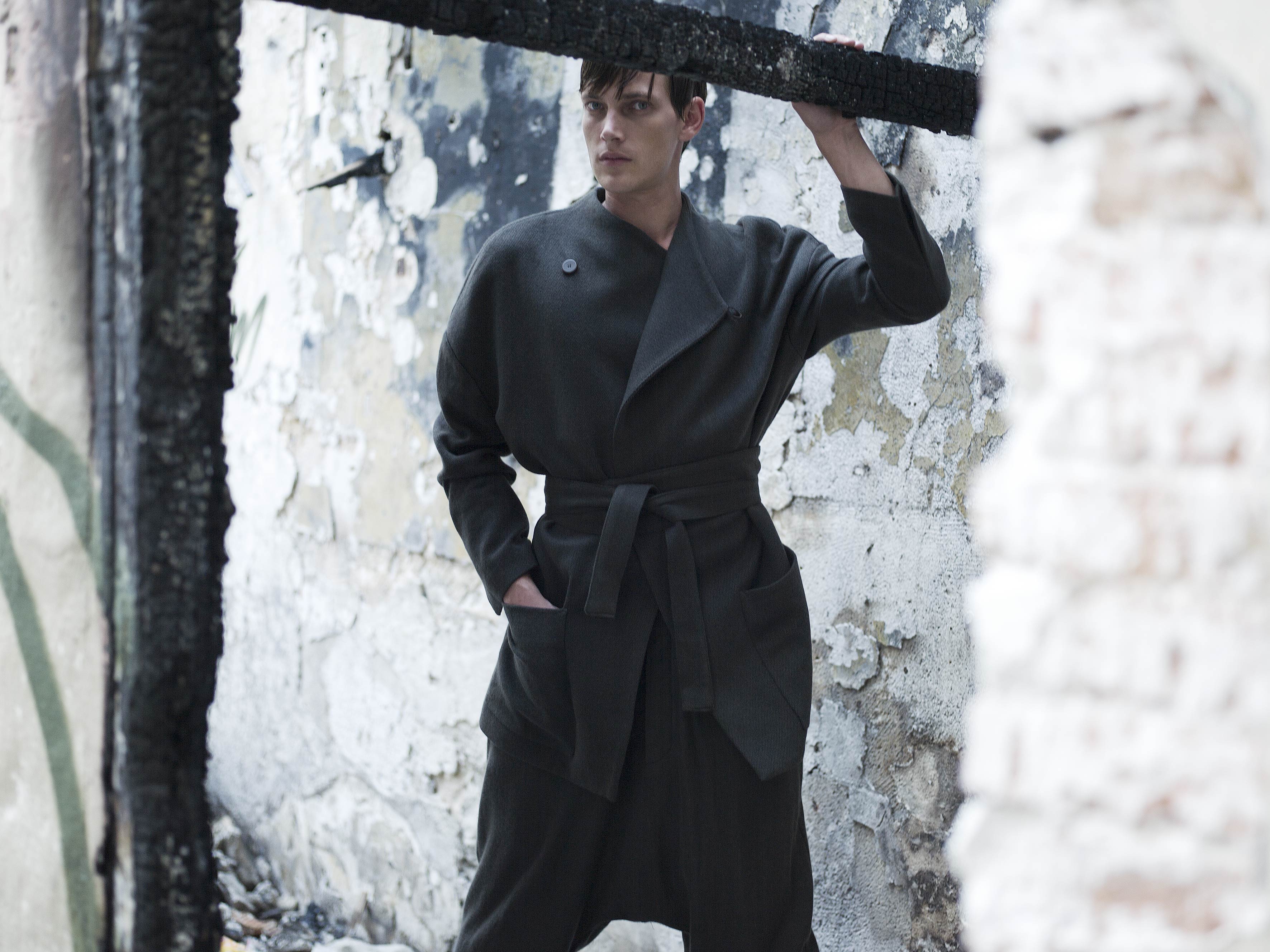 Person in a stylish dark robe and pants stands in a weathered, urban setting with peeling walls, surrounded by a rough, industrial frame. They gaze to the side, exuding a sense of confidence and contemplation.