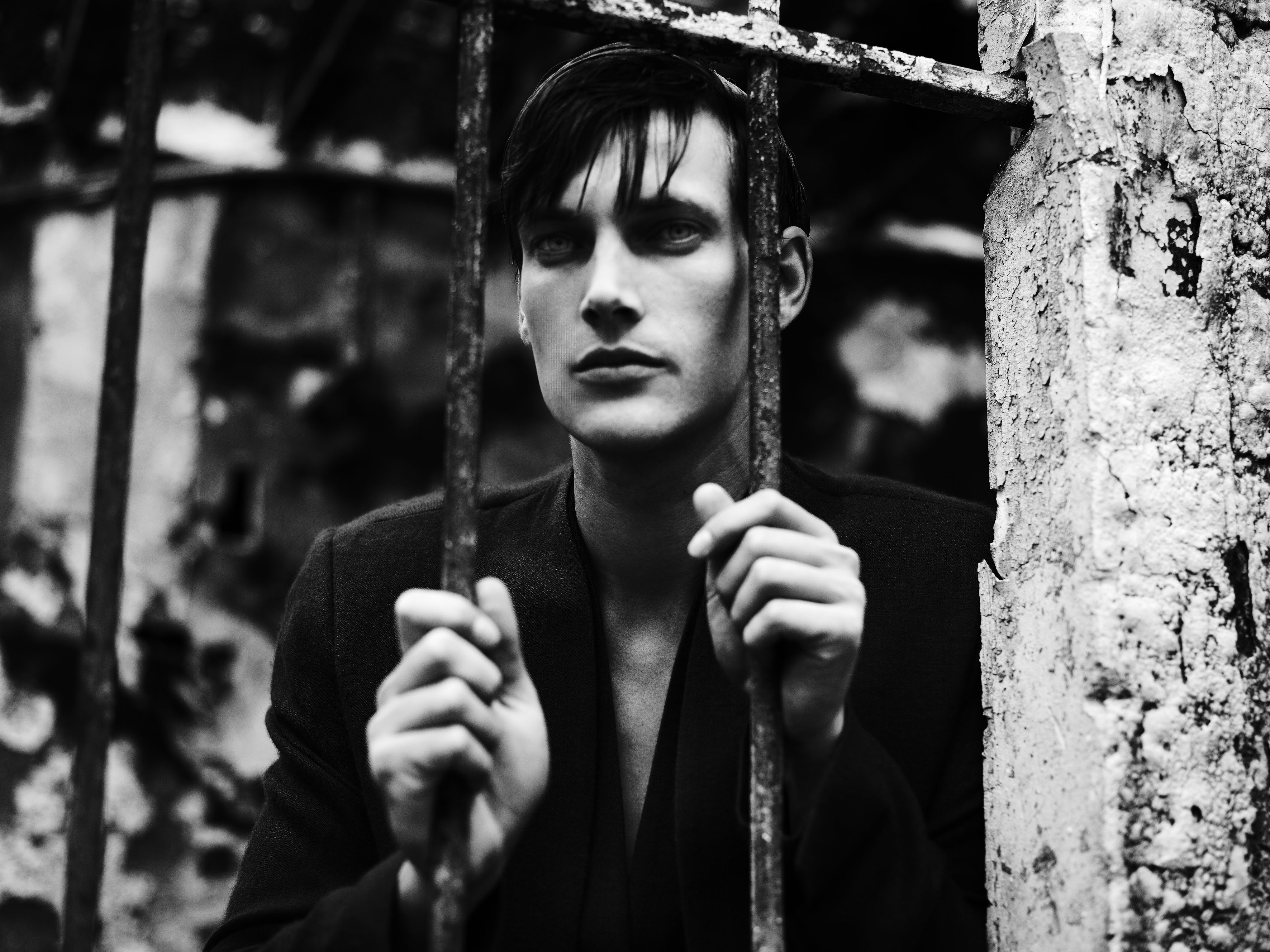 Black and white image of a person with short, dark hair, standing behind metal bars, holding onto them with both hands. The background is blurred, showing a weathered wall. The mood appears somber or contemplative.