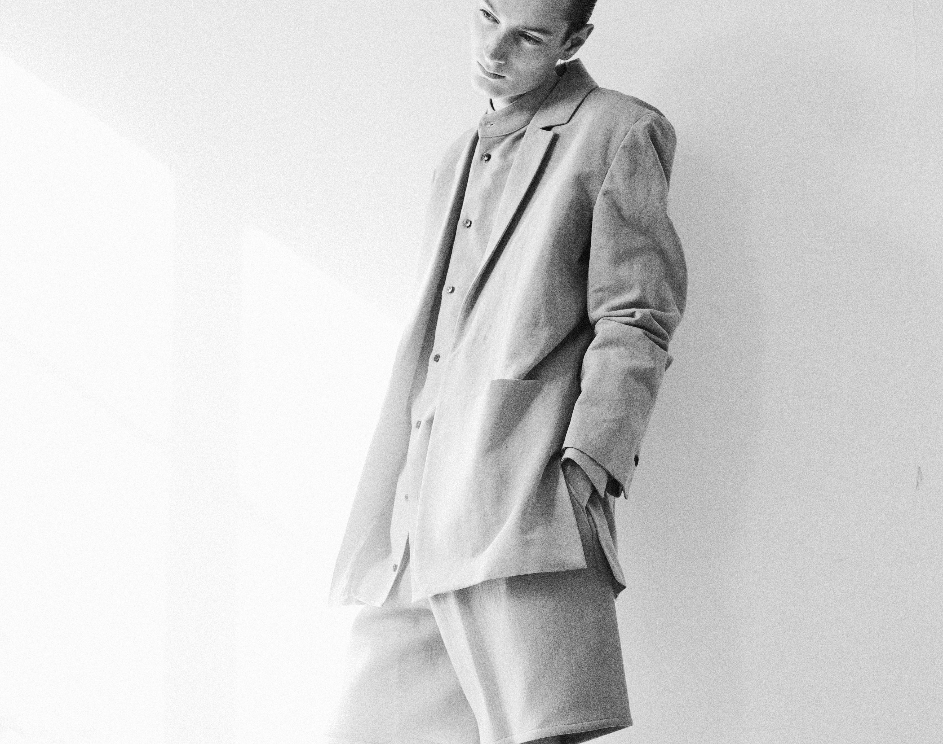 A person in grayscale wearing a loose-fitting suit and shorts leans against a wall. The soft lighting creates a relaxed and stylish atmosphere.