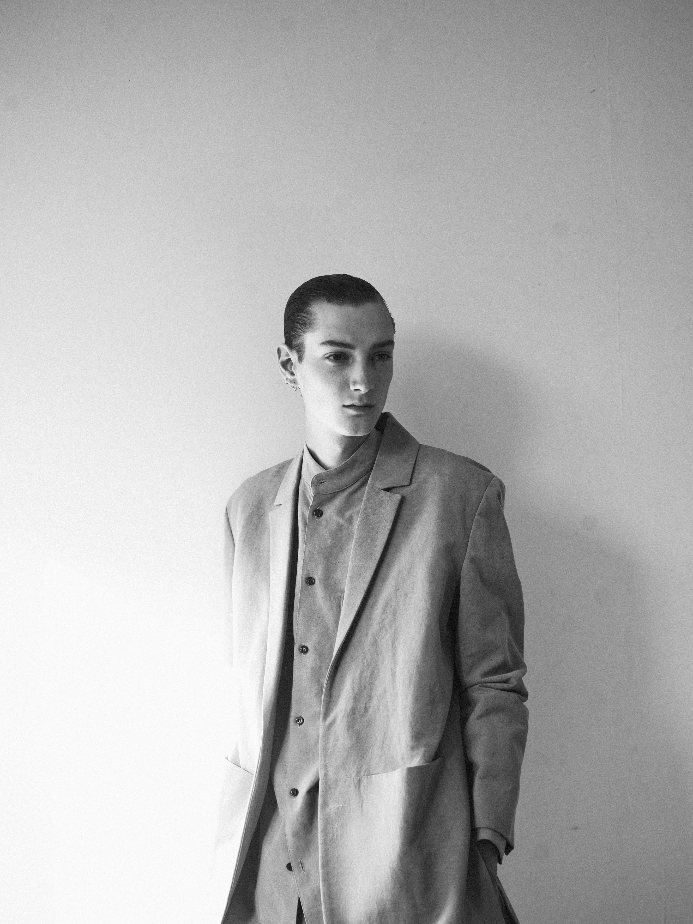 A black and white photo of a person wearing a tailored coat and shirt, standing against a plain wall. The lighting creates soft shadows, adding depth to the image. The persons hair is styled neatly, and they look slightly away from the camera.