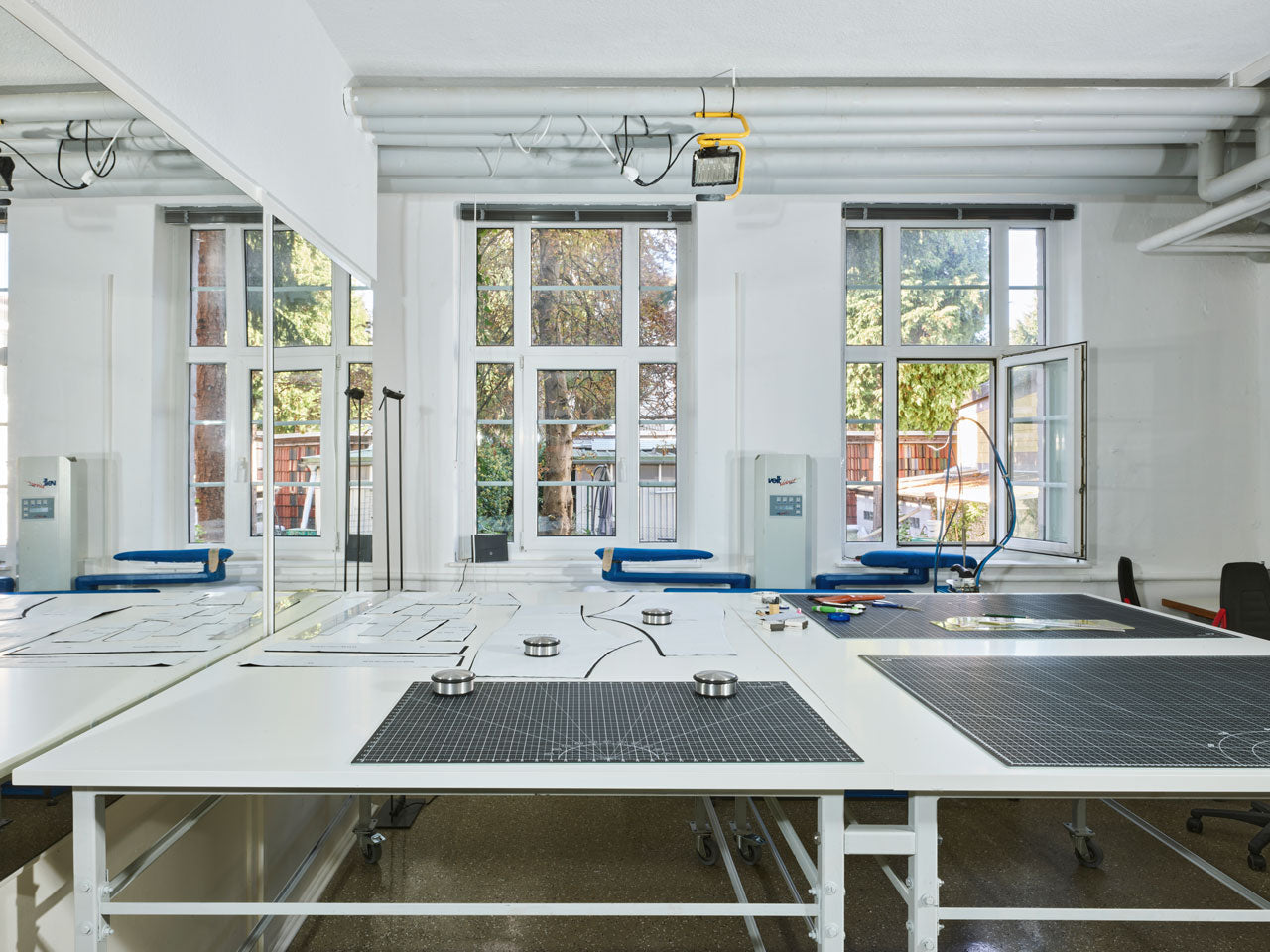 A bright sewing room with large windows and drafting tables covered in patterns and tools. The room has white walls, mirrors, and blue chairs. An open window lets in natural light, and trees are visible outside.