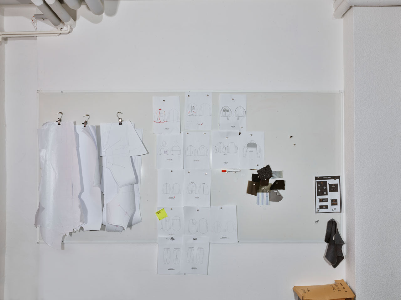 A whiteboard with numerous fashion design sketches and fabric swatches pinned to it. There are several paper patterns hanging, and a small section of electronic controls is visible on the right side.