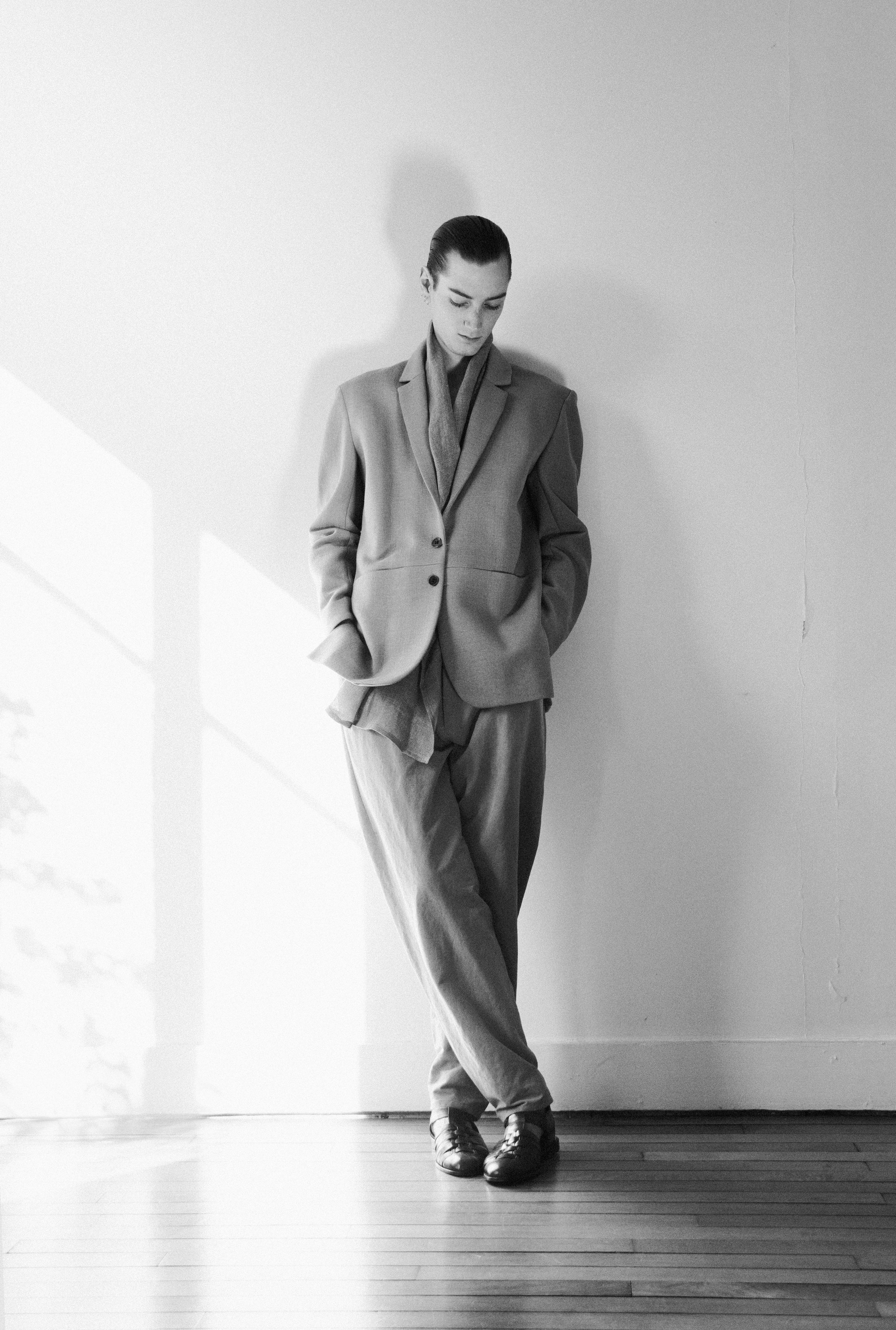 Black and white image of a person standing against a wall in a relaxed pose, wearing a tailored suit with hands in pockets. Soft natural light casts shadows on the wall, creating a calm, elegant atmosphere.