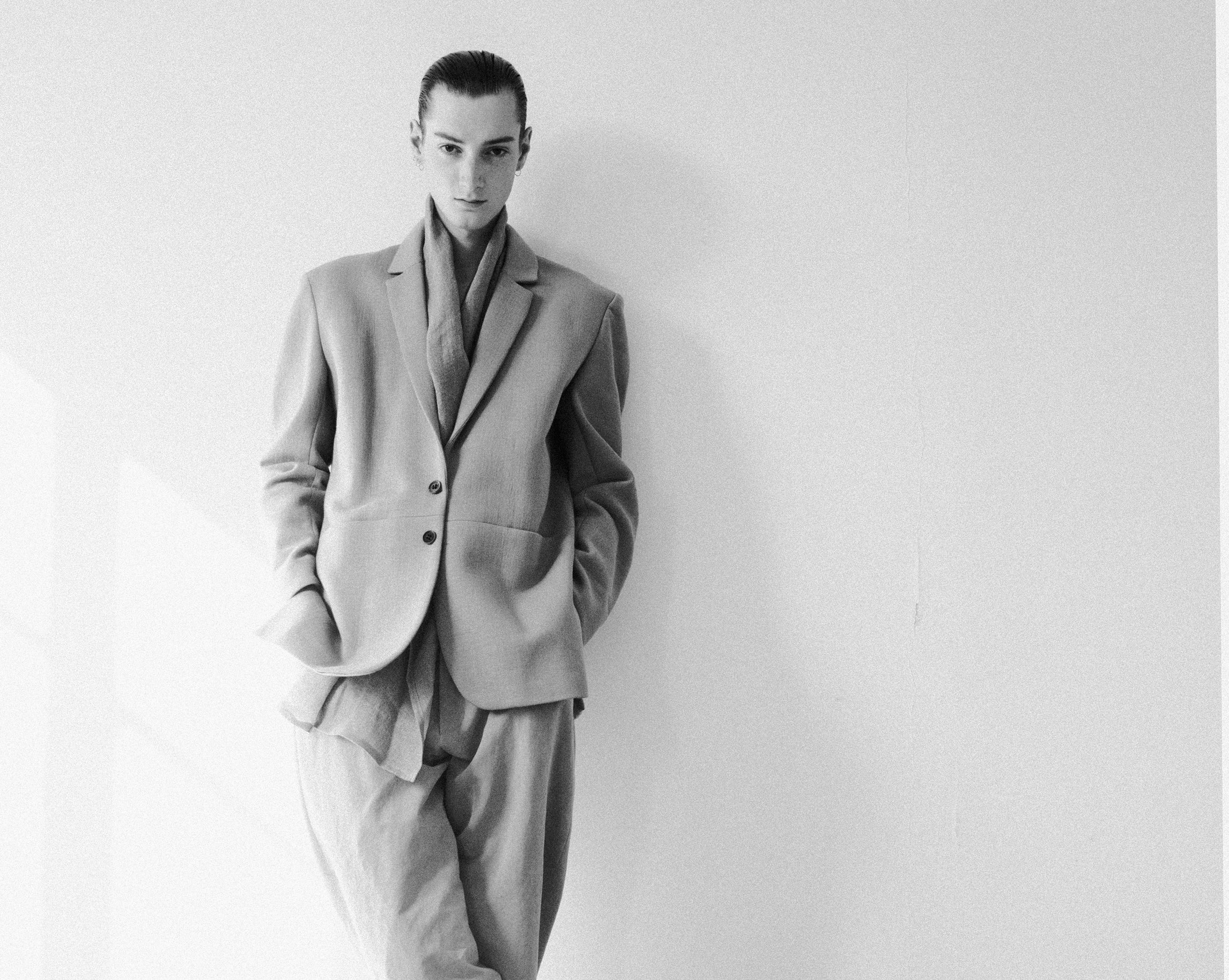 A person stands against a white wall in a black and white photo, wearing an oversized suit with hands in pockets. The natural light casts gentle shadows, creating a minimalist and elegant atmosphere.