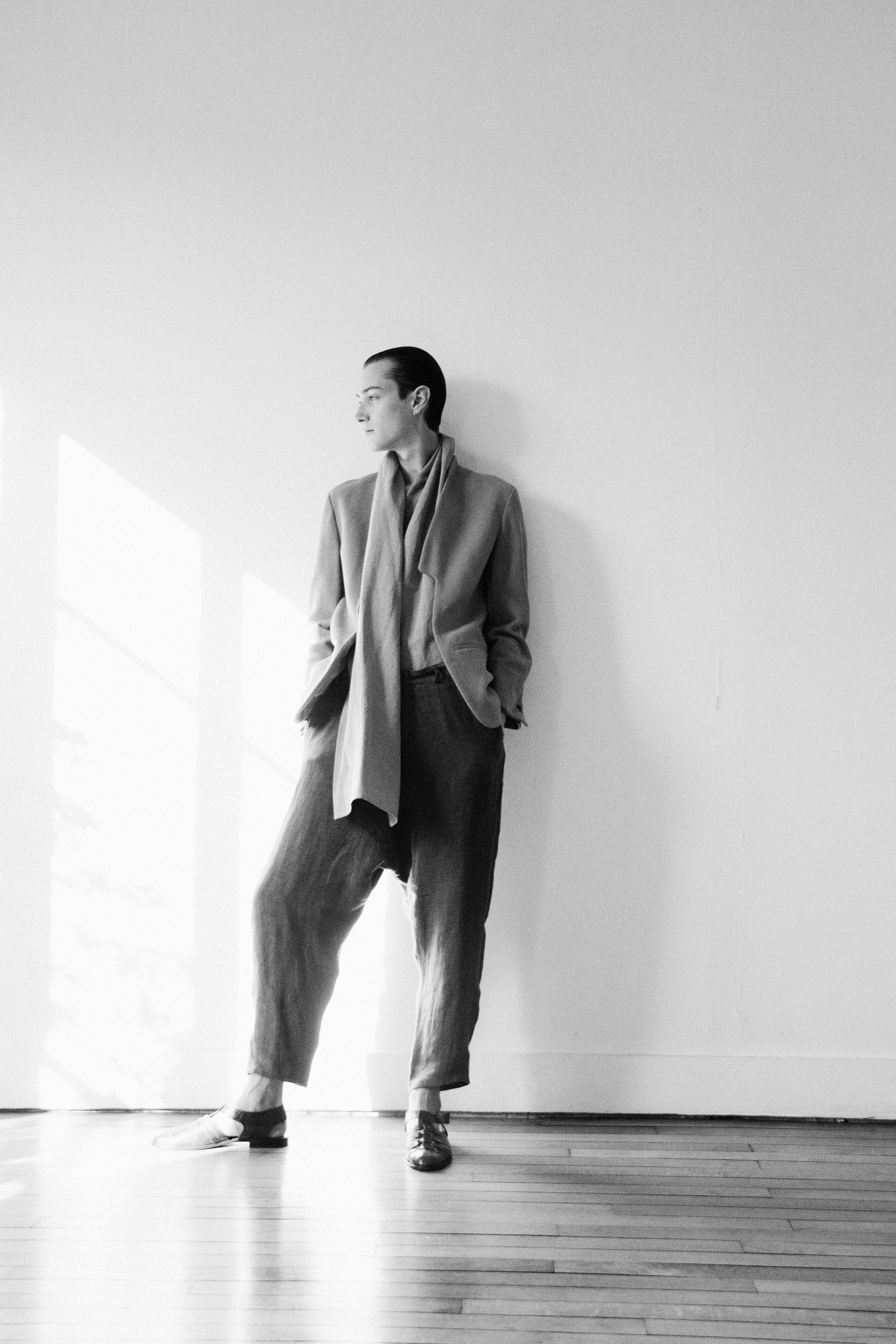 A person with short hair leans against a wall in a bright room, wearing a loose-fitting suit and looking to the side. Sunlight streams through windows, casting shadows on the floor. The image is in black and white.