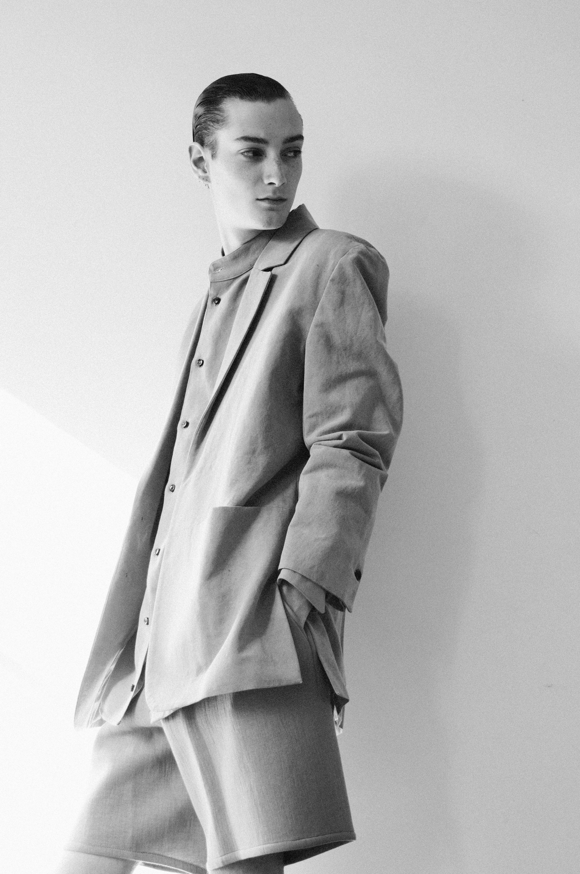 Black and white photo of a person with short hair, wearing an oversized blazer and shorts. They pose confidently, looking to the side, with one hand in a pocket. The background is a plain wall, creating a minimalist, stylish ambiance.