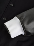 A close-up of the "Silverskin" College Jacket by gabrielstunz highlights the European craftsmanship featuring a black body, black faux leather sleeve, and white ribbed cuff. It showcases smooth fabric with visible seam stitching and two partially visible black snap buttons.