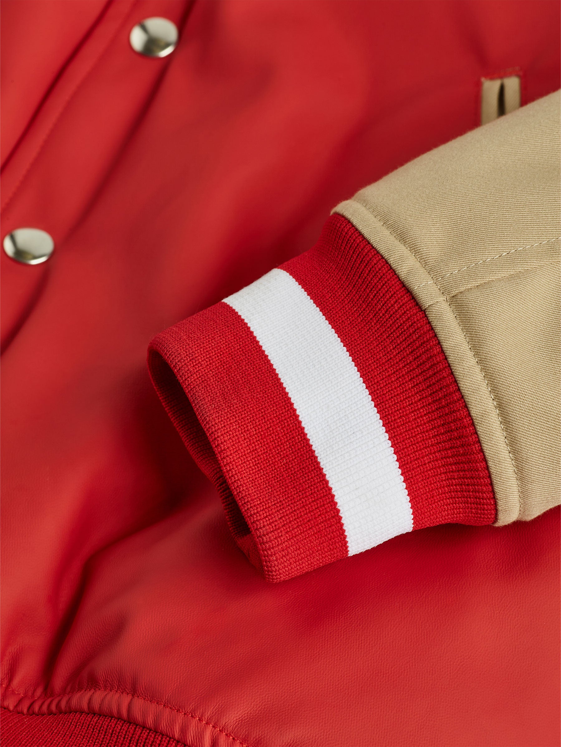 A close-up of the gabrielstunz Red College Jacket with Beige Twill Sleeves highlights its classic collegiate style. The beige sleeve is complemented by a red and white ribbed cuff, while the body features premium materials and silver snap buttons. Together, these textures and colors embody a timeless, sporty look.