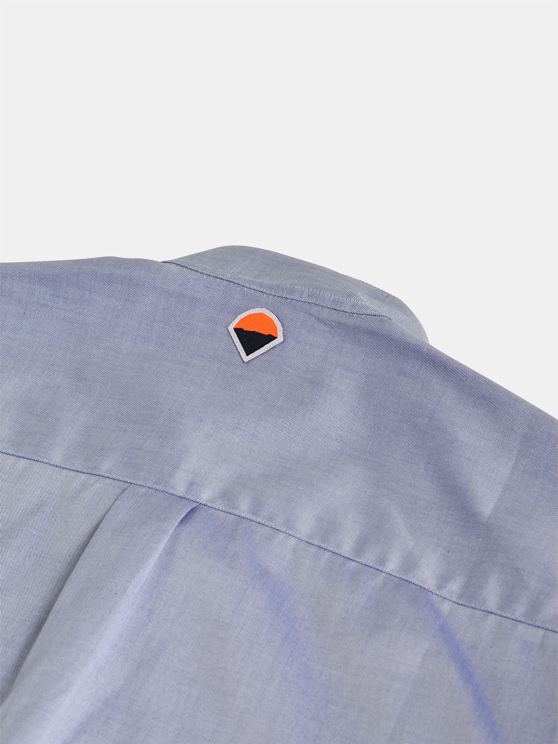 Close-up of the back of a light blue POCKETPARACHUTE® with Mao Collar shirt by gabrielstunz, showcasing a small triangular logo embroidered near the neckline in black, orange, and white colors. The premium fabric has a smooth texture that enhances the elegance of this design.