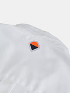 Featuring a close-up of a white garment, the gabrielstunz POCKETPARACHUTE® Shirt highlights a small embroidered logo with an eye-catching orange and black abstract design. The premium fabric looks smooth and lightweight against a plain background, emphasizing its modern fit.