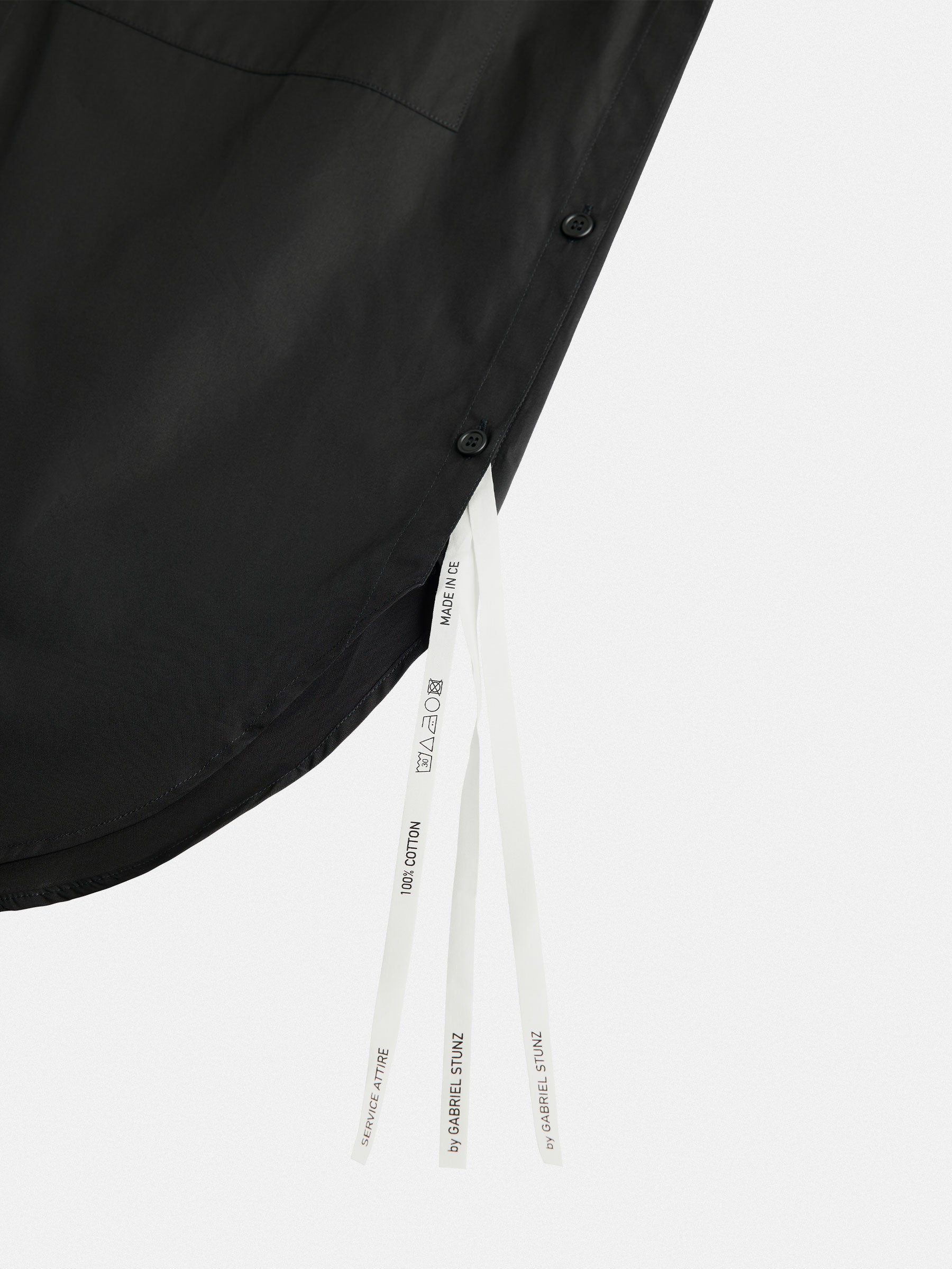 A close-up of the hem on the Oversized Cotton Shirt by gabrielstunz reveals two white tags highlighting its premium cotton and care instructions. The shirt boasts an oversize fit with side buttons, embodying European craftsmanship through its minimalist and modern design against a plain background.