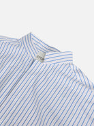 Close-up of the Oversized Mandarin Collar Shirt by gabrielstunz, showcasing its white fabric adorned with blue vertical stripes crafted from 100% cotton by Cottonificio Albini. The shirt is displayed on a flat surface, revealing a visible tag inside the collar reading STU.