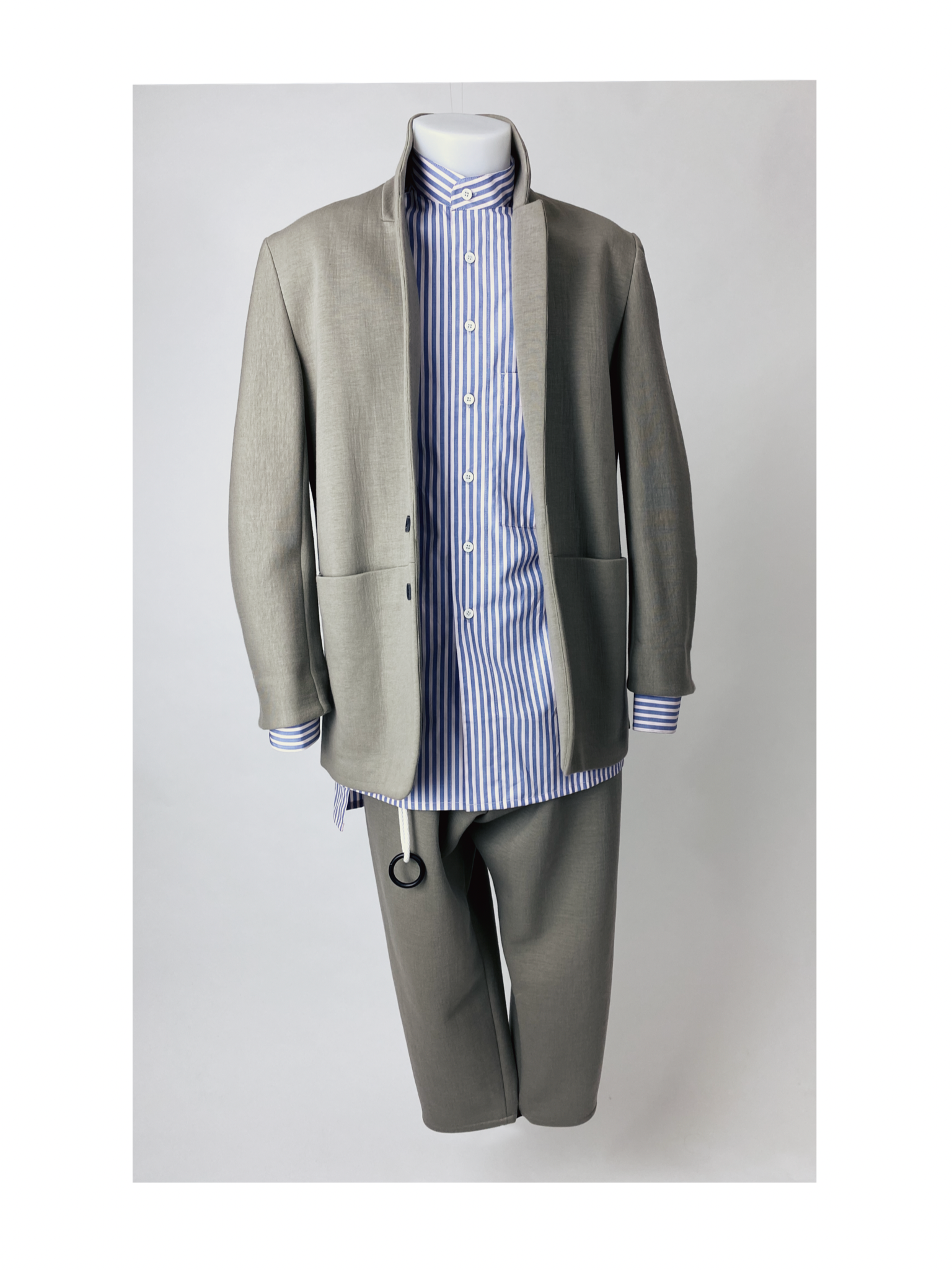 A mannequin wearing a light gray suit with a standing collar jacket, over a blue and white striped shirt. The suit includes matching trousers, and theres a black ring accessory hanging from the pocket.