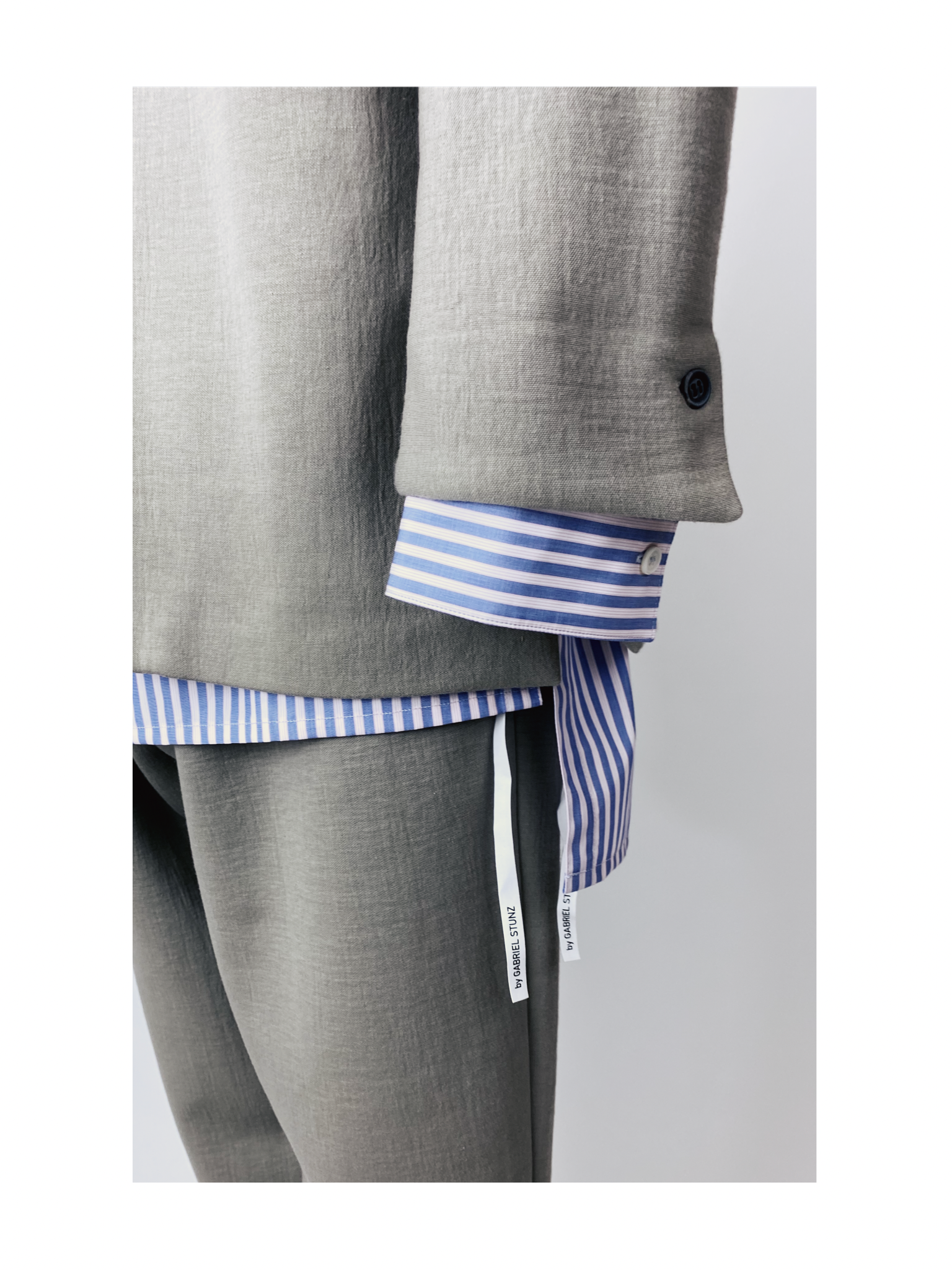 Close-up of a person wearing a light gray suit with a blue and white striped shirt underneath. The shirt cuffs extend beyond the suit sleeves, showing button details. White tags with text hang from the shirt.