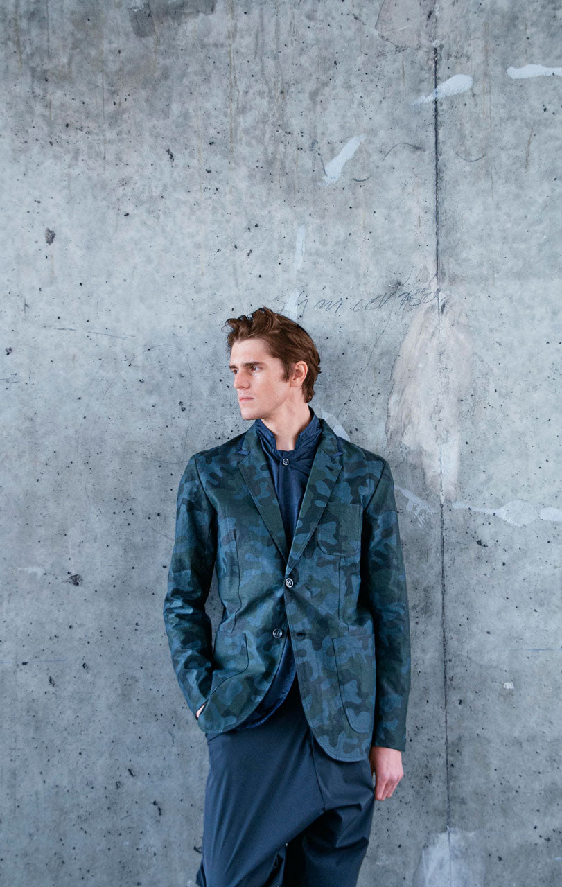 A person wearing a blue camouflage suit jacket and dark trousers stands against a textured gray concrete wall. They have one hand in their pocket and are looking to the side.