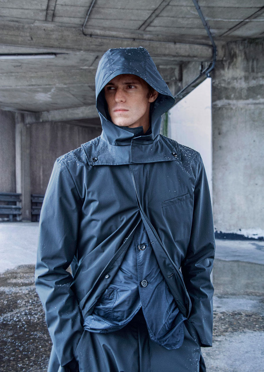 A person wearing a dark raincoat with a hood stands under a concrete structure. Water droplets are visible on the coat. The background shows an urban environment with grey walls and a wet pavement.