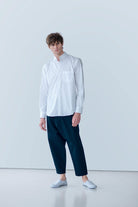 A person stands against a light gray background wearing a white POCKETPARACHUTE® Shirt by gabrielstunz, crafted from premium fabric, paired with dark blue pants and white slip-on shoes. Their hair is styled casually, and they exhibit a relaxed posture.