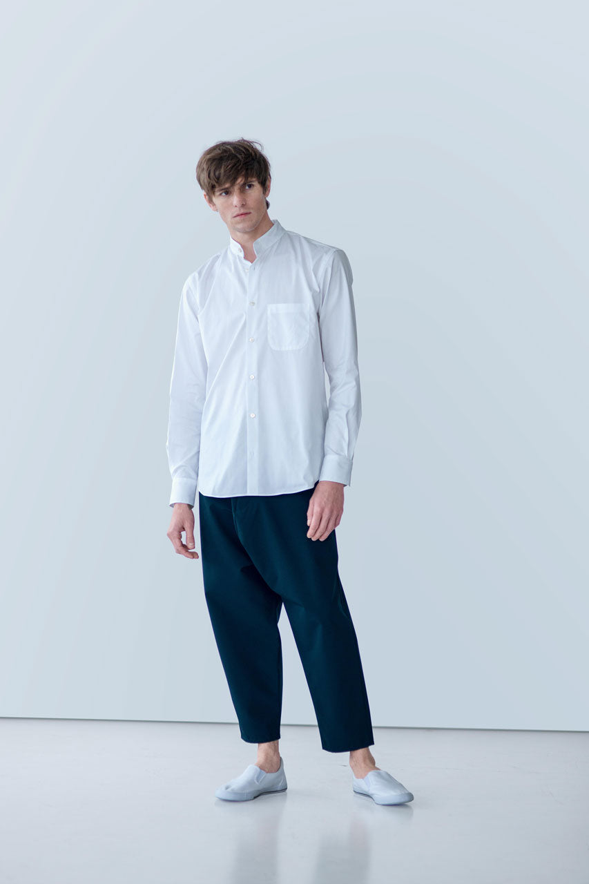 A person stands against a light gray background wearing a white POCKETPARACHUTE® Shirt by gabrielstunz, crafted from premium fabric, paired with dark blue pants and white slip-on shoes. Their hair is styled casually, and they exhibit a relaxed posture.