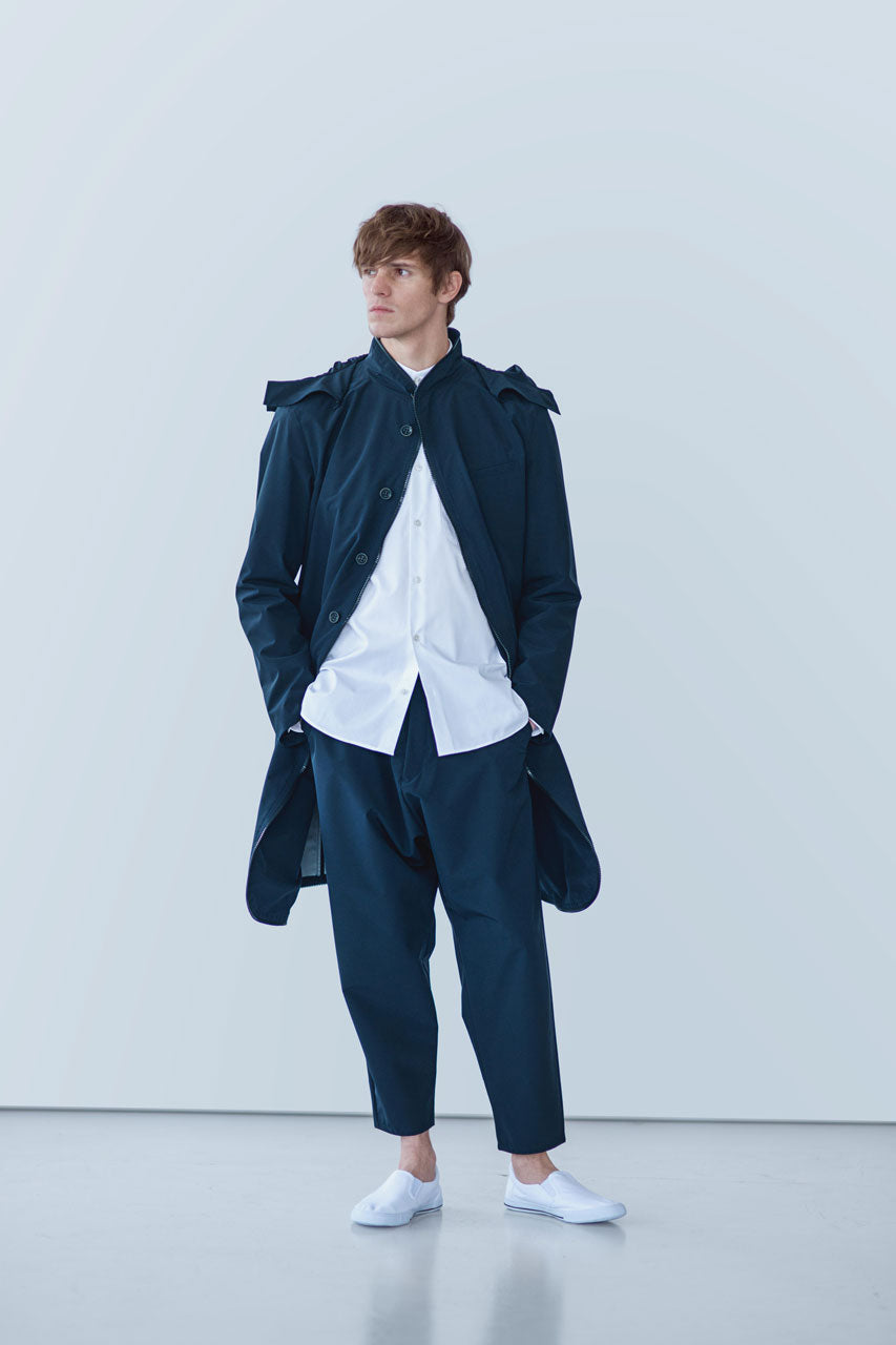 A person stands wearing a gabrielstunz POCKETPARACHUTE® Raincoat in navy blue, merging packable convenience with style, over a white shirt and matching pants, paired with water-repellent slip-on shoes. The background is a light gray wall, and the person's gaze is directed to the side.