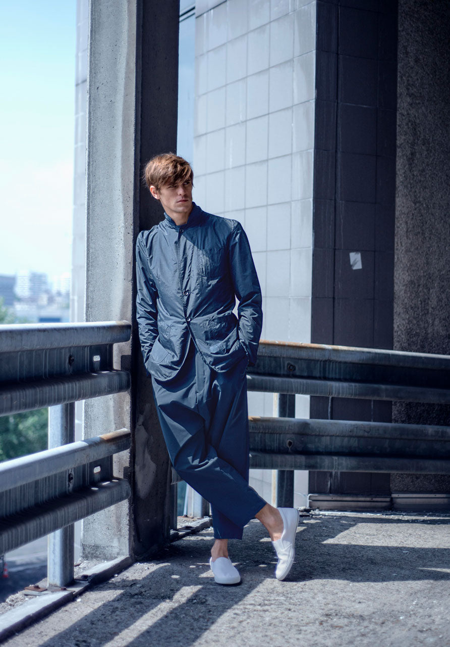 A person in a blue outfit stands casually against a railing in an urban setting. They are wearing white slip-on shoes. The background features concrete walls and a distant view of the city skyline.