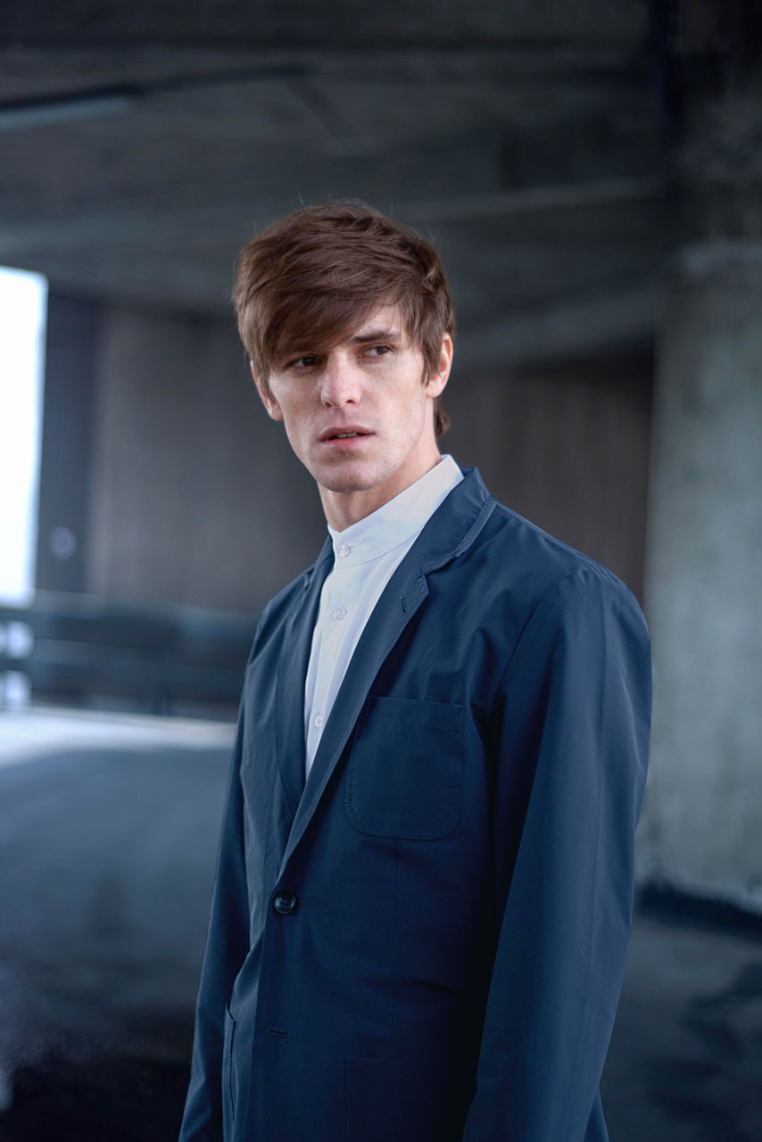 A man with brown hair wearing a blue suit and white shirt stands in an urban, partially lit setting with concrete walls. He gazes into the distance with a serious expression.