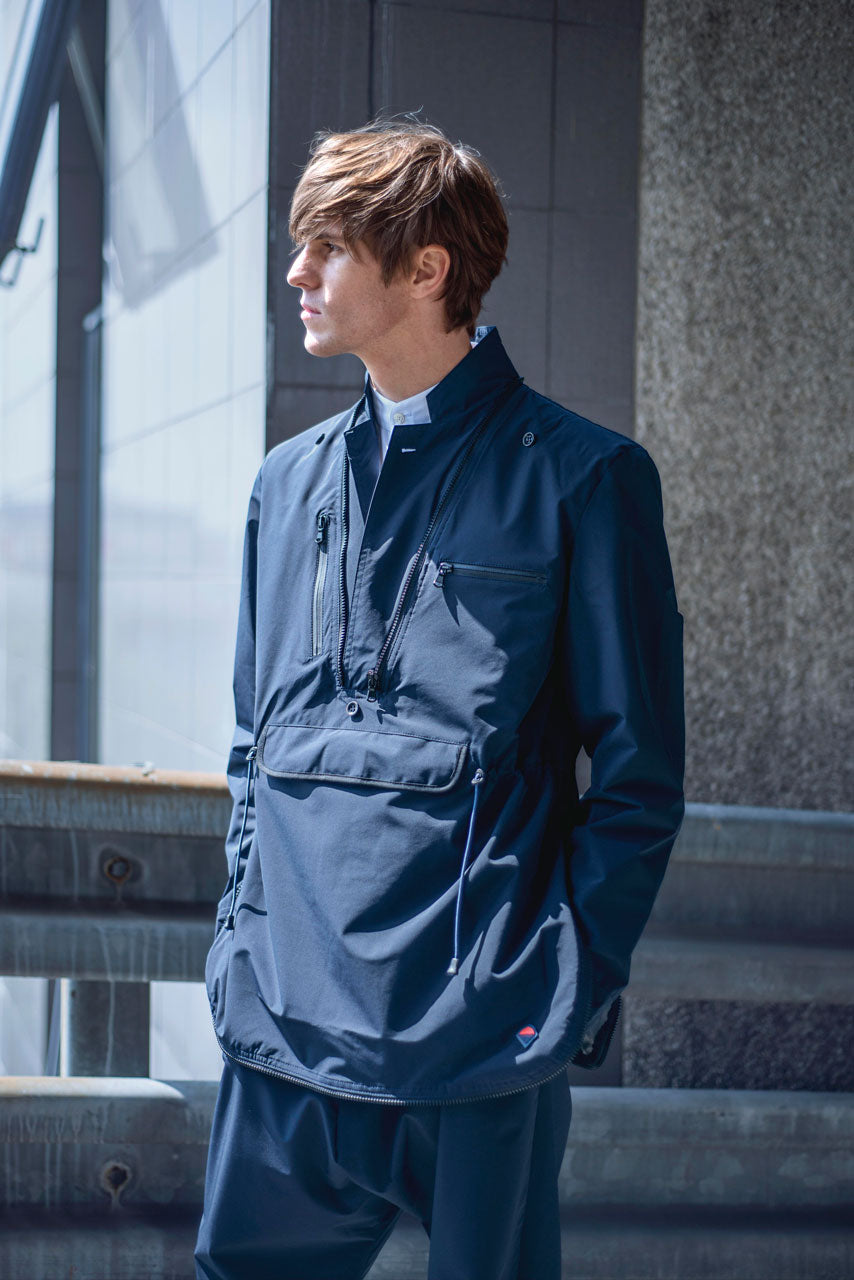 A person wearing a dark blue, long-sleeved jacket with multiple pockets stands beside a railing. The individual has short brown hair and is gazing to the side. The background features a large window and a textured wall.