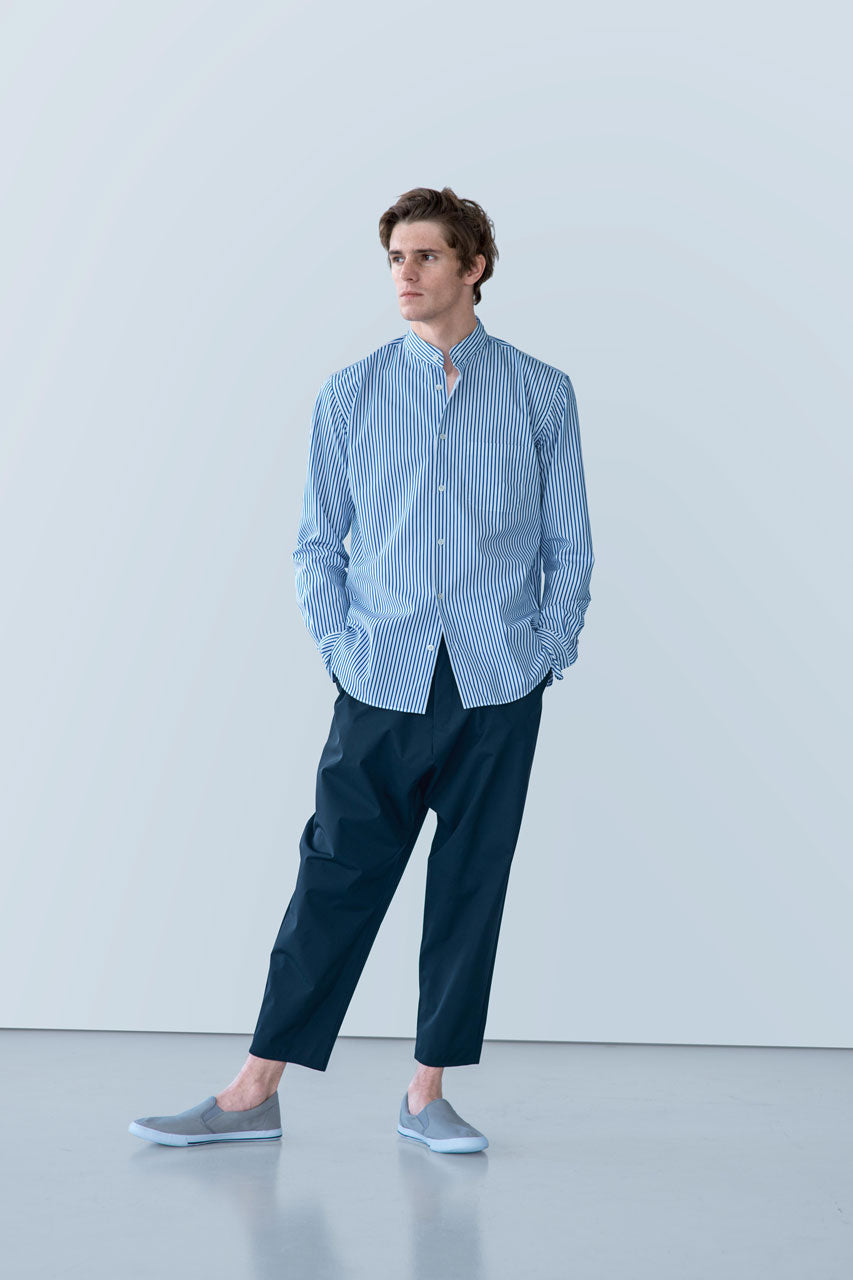 A man stands against a light gray background wearing a blue and white striped gabrielstunz POCKETPARACHUTE® Shirt with Mao Collar, featuring mother-of-pearl buttons. He pairs it with dark blue pants and light blue slip-on shoes, gazing to the side with his hands in his pockets.