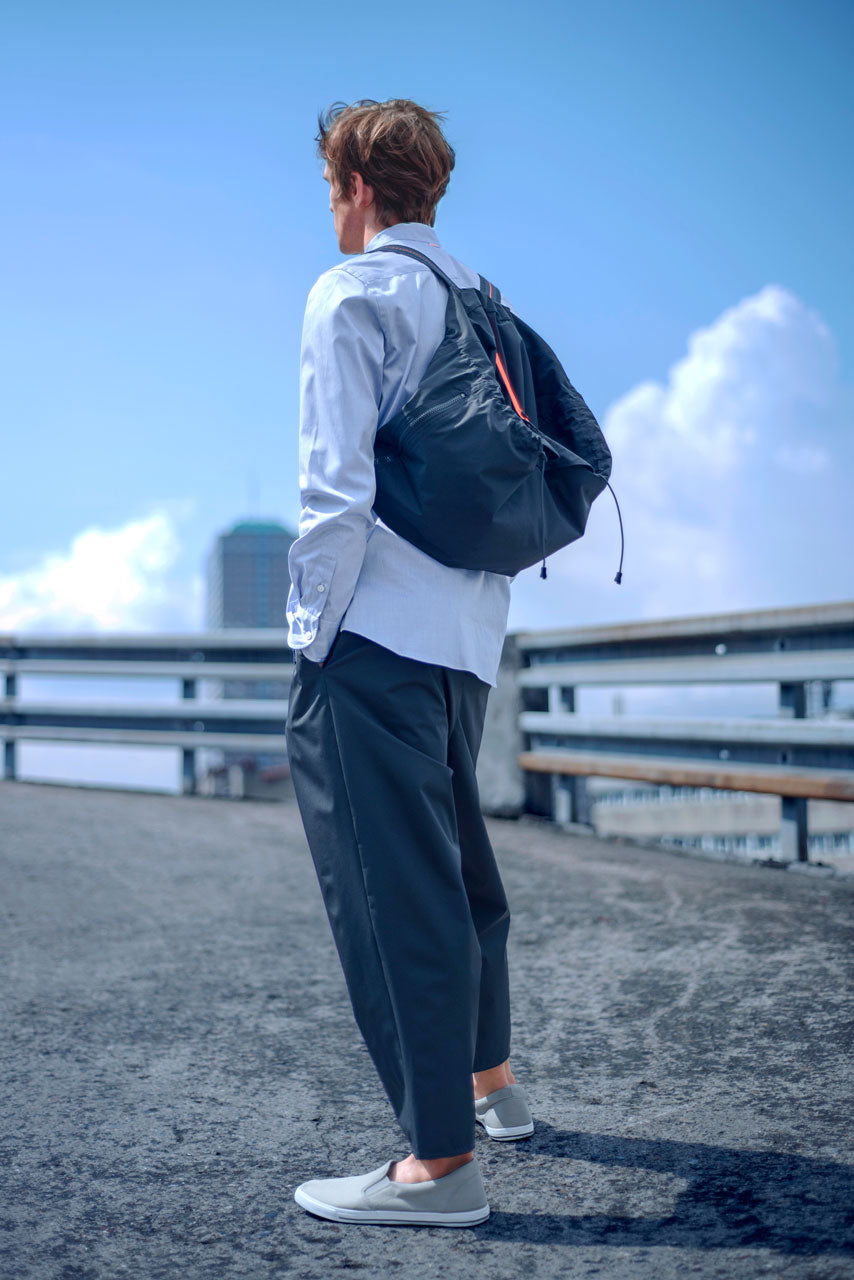 A person with short hair stands on a rooftop, wearing a light blue long-sleeve shirt, dark pants, and gray slip-on shoes. They have a black drawstring backpack and are looking towards the city skyline under a bright blue sky with clouds.