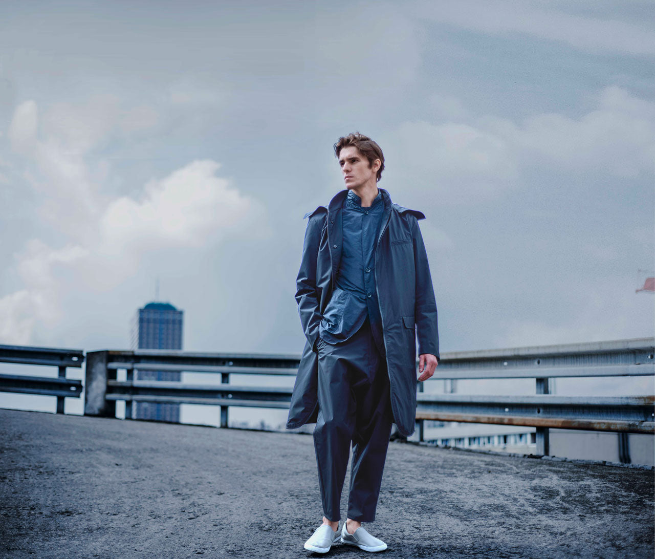 A person stands on a rooftop, wearing a long, dark coat and blue clothing underneath. The sky is cloudy, and a tall building is visible in the background. The person is looking to the side, with hands in pockets and wearing white shoes.