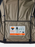 The image showcases the inside back panel of a khaki POCKETPARACHUTE Jacket from gabrielstunz, featuring a large label with an orange parachute design. It includes diagram instructions for The Original POCKETPARACHUTE® feature, emphasizing its packable convenience. Black strings can be seen on the sides.