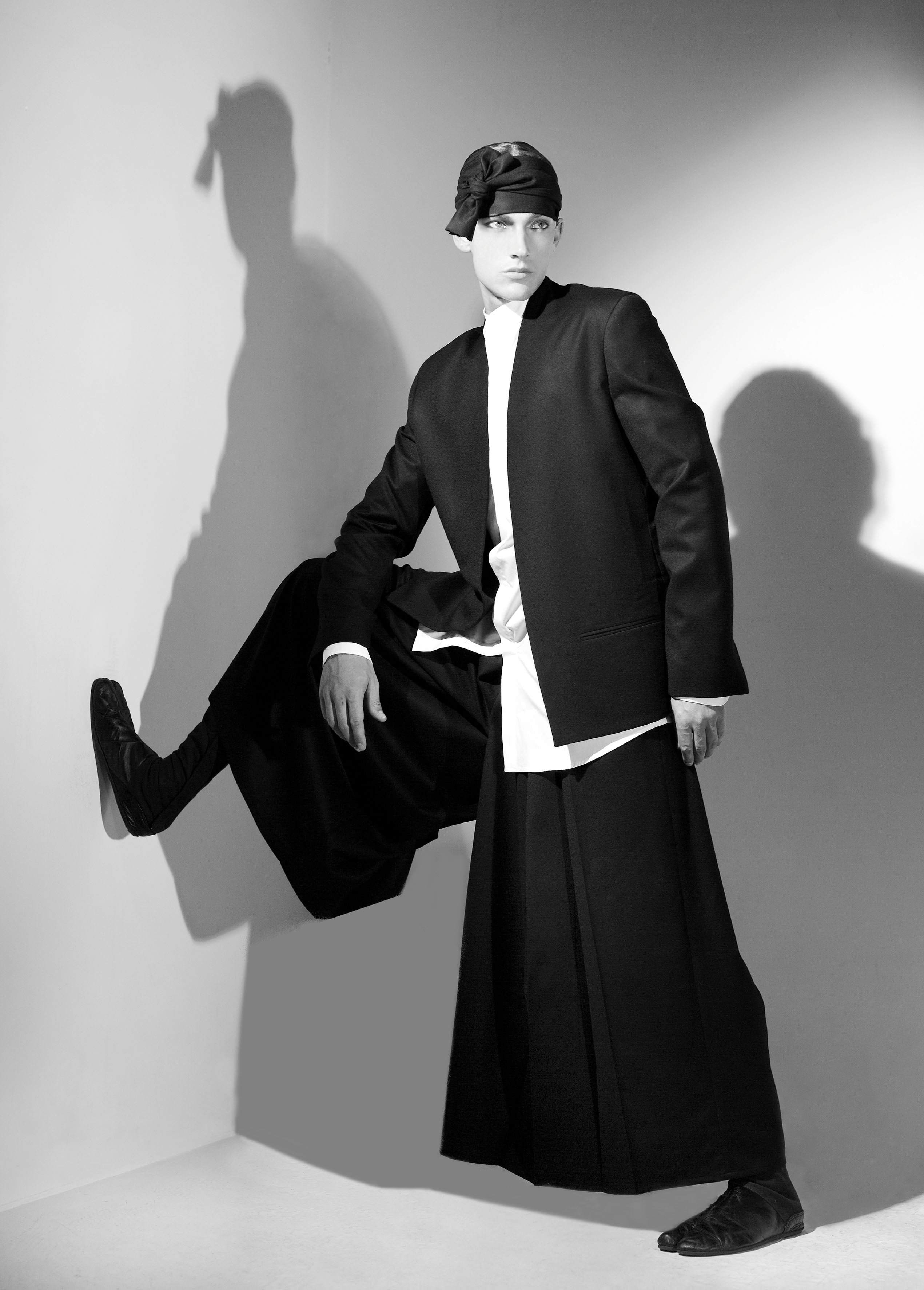 A person in a monochrome outfit poses dramatically against a wall. They wear a black jacket, long black skirt, white shirt, and black cap. One leg is bent with the foot resting on the wall, casting a shadow in the soft lighting.