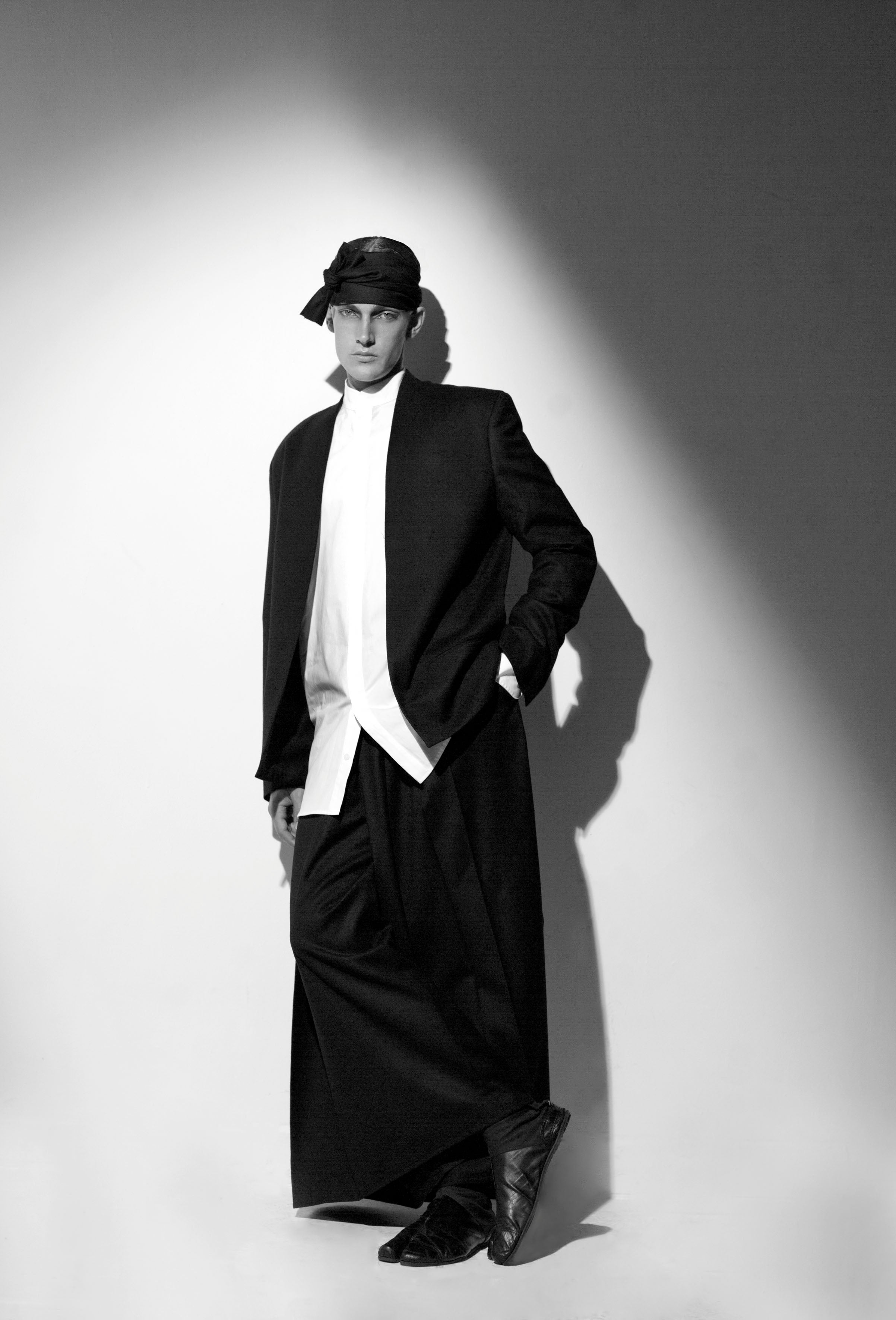 A person in a stylish outfit poses against a wall, with dramatic lighting casting shadows. They wear a black hat, long black coat, white shirt, wide black trousers, and black shoes, creating a bold, monochromatic look.