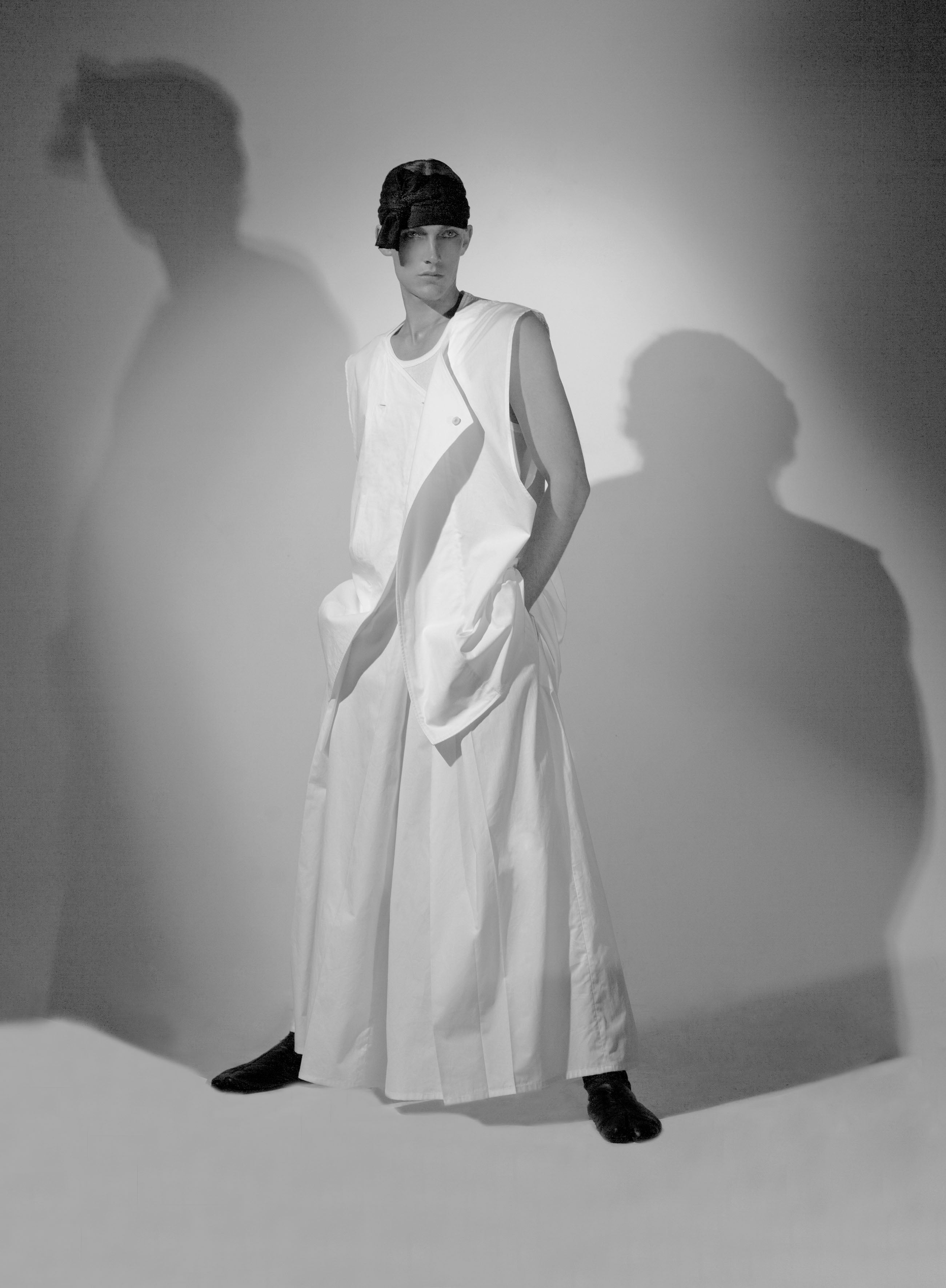 A person wearing a long, flowing white outfit and a dark cap stands confidently with hands in pockets. The background is lit to create dramatic shadows, enhancing the artistic and fashion-forward mood.