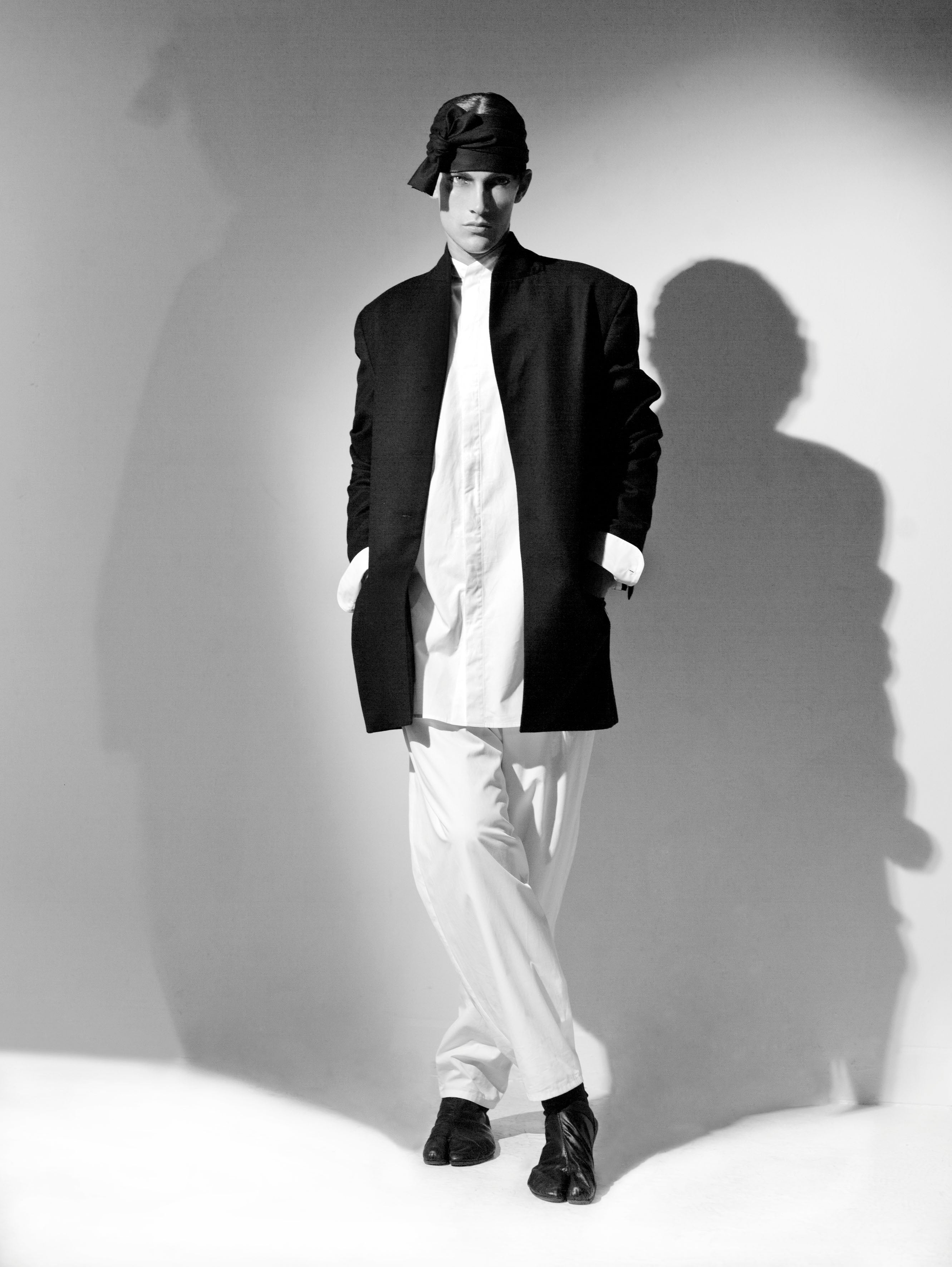 Black and white photo of a person standing against a wall, dressed in a long black coat, white shirt, and light pants. They wear a dark headwrap and boots, with hands in pockets. Theres a shadow cast on the wall behind them.