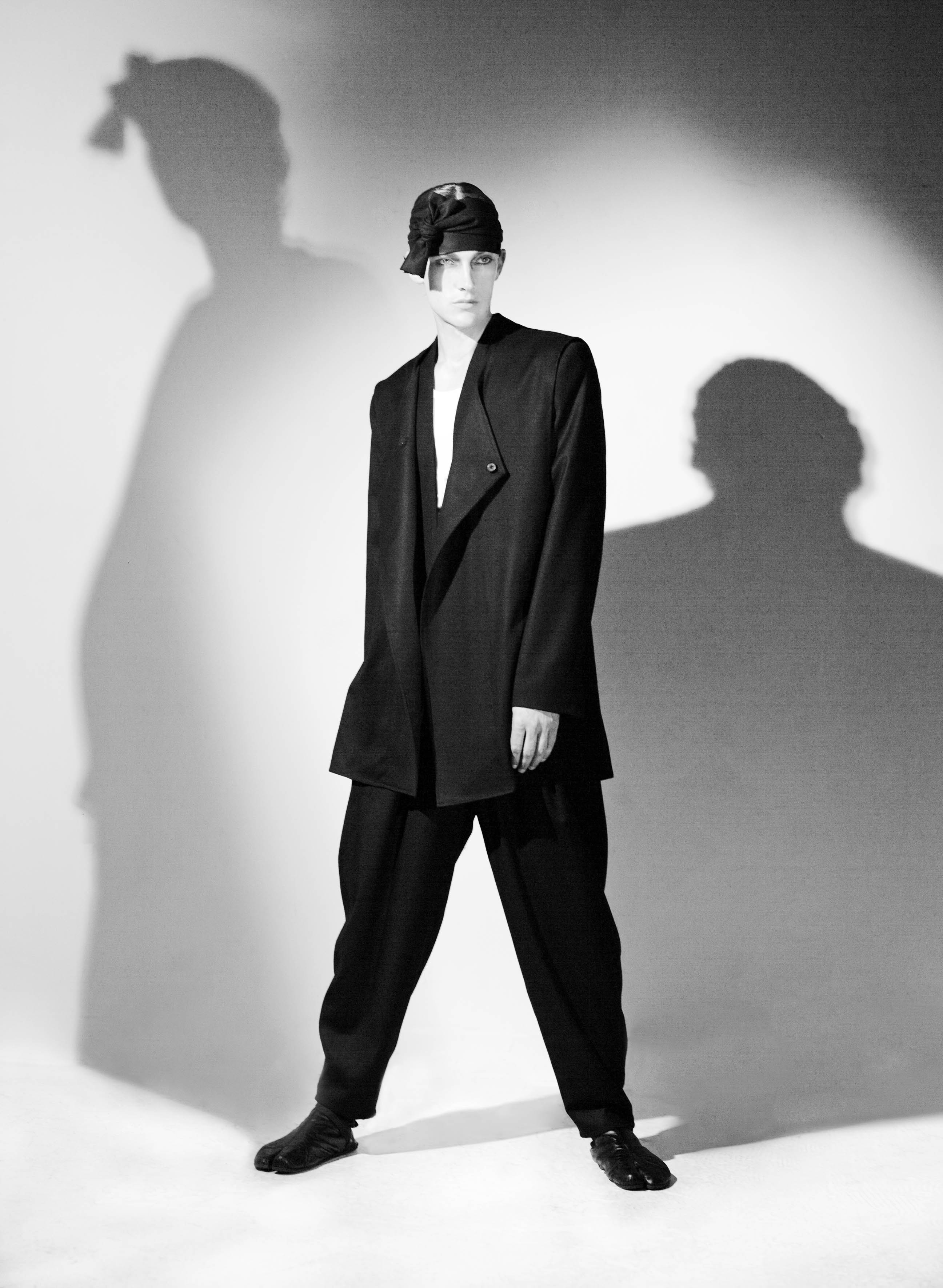 A person stands confidently in a black oversized suit and hat against a plain backdrop. The lighting creates a dramatic shadow on the wall. The image is in black and white, adding a classic, artistic feel.