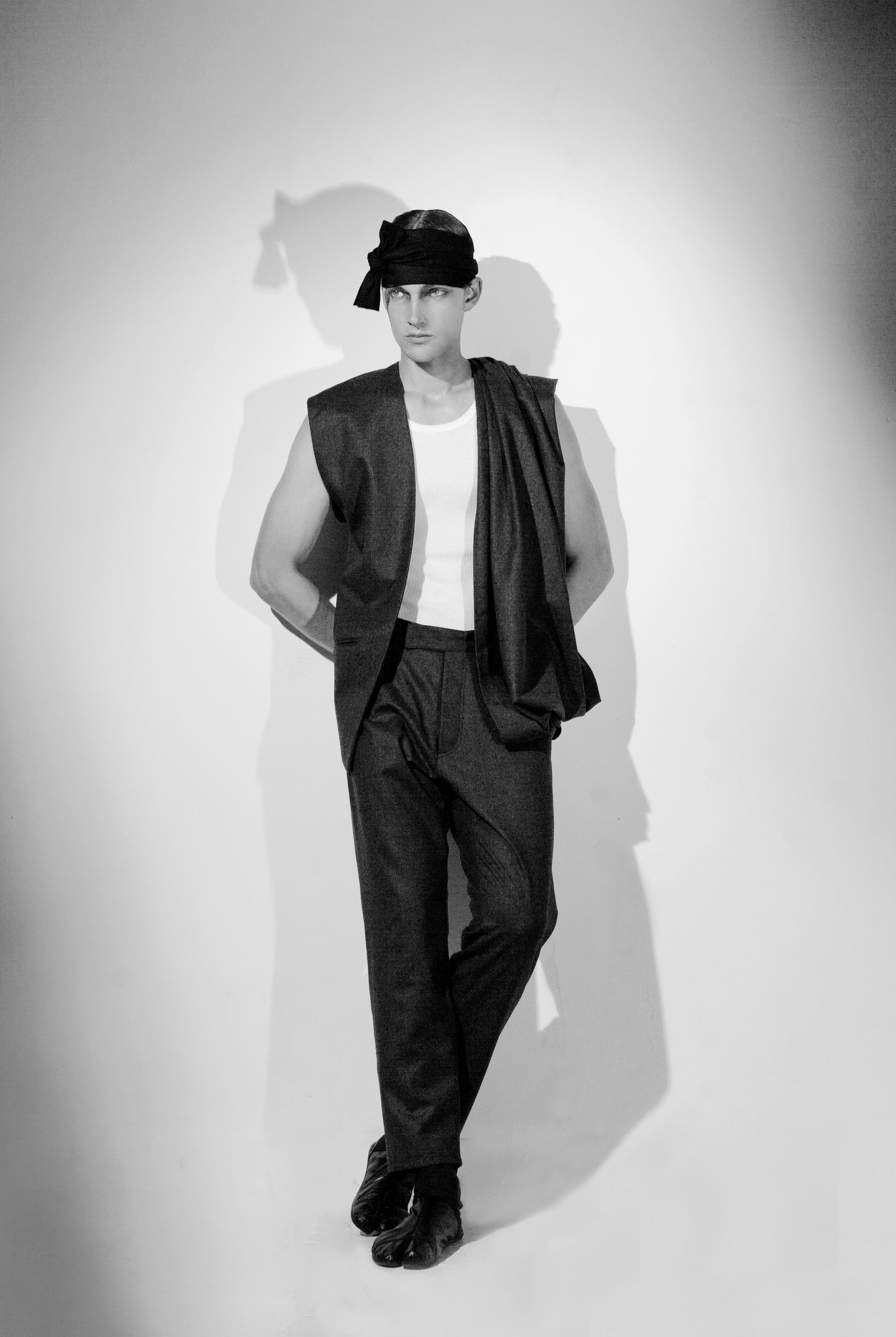 A person stands against a plain backdrop wearing a hat, sleeveless coat, white shirt, and dark pants with hands behind their back. The image is in black and white with dramatic lighting creating a shadow on the wall.