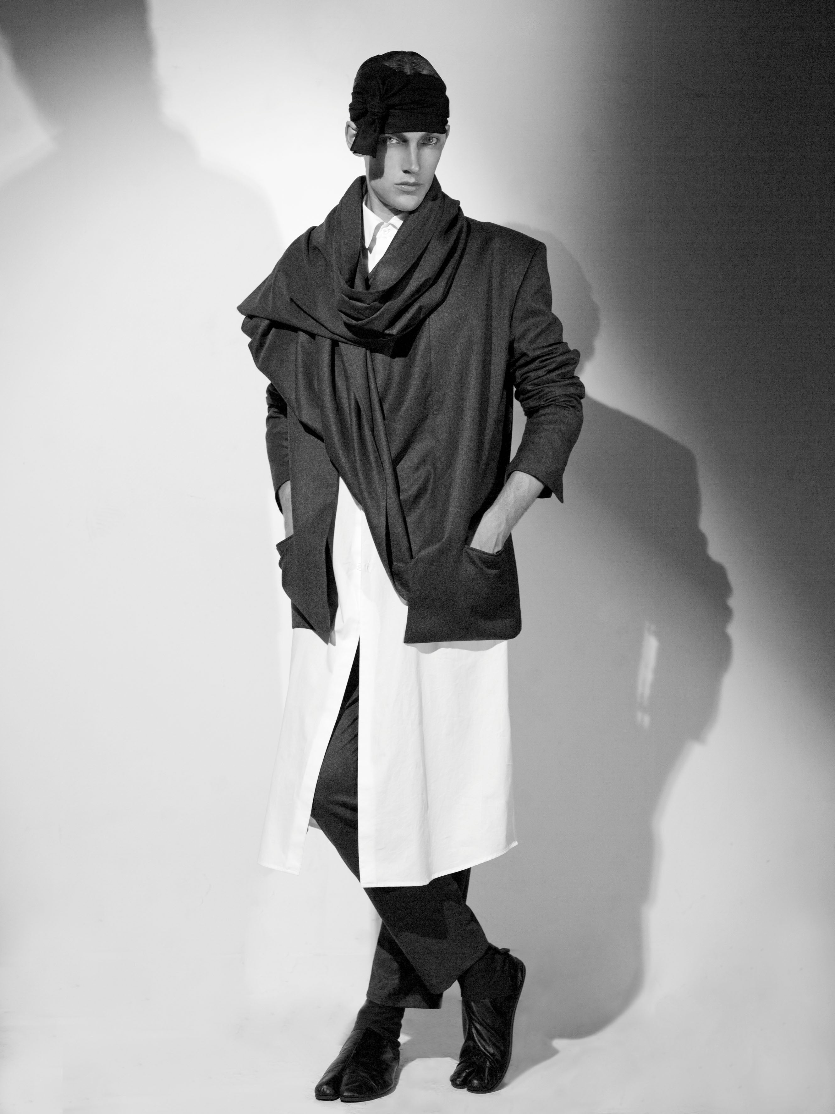 Black and white photo of a fashion model wearing a layered outfit, including a draped scarf, long white shirt, tailored dark jacket, and matching pants. They are posing with hands in pockets against a shadowed wall.