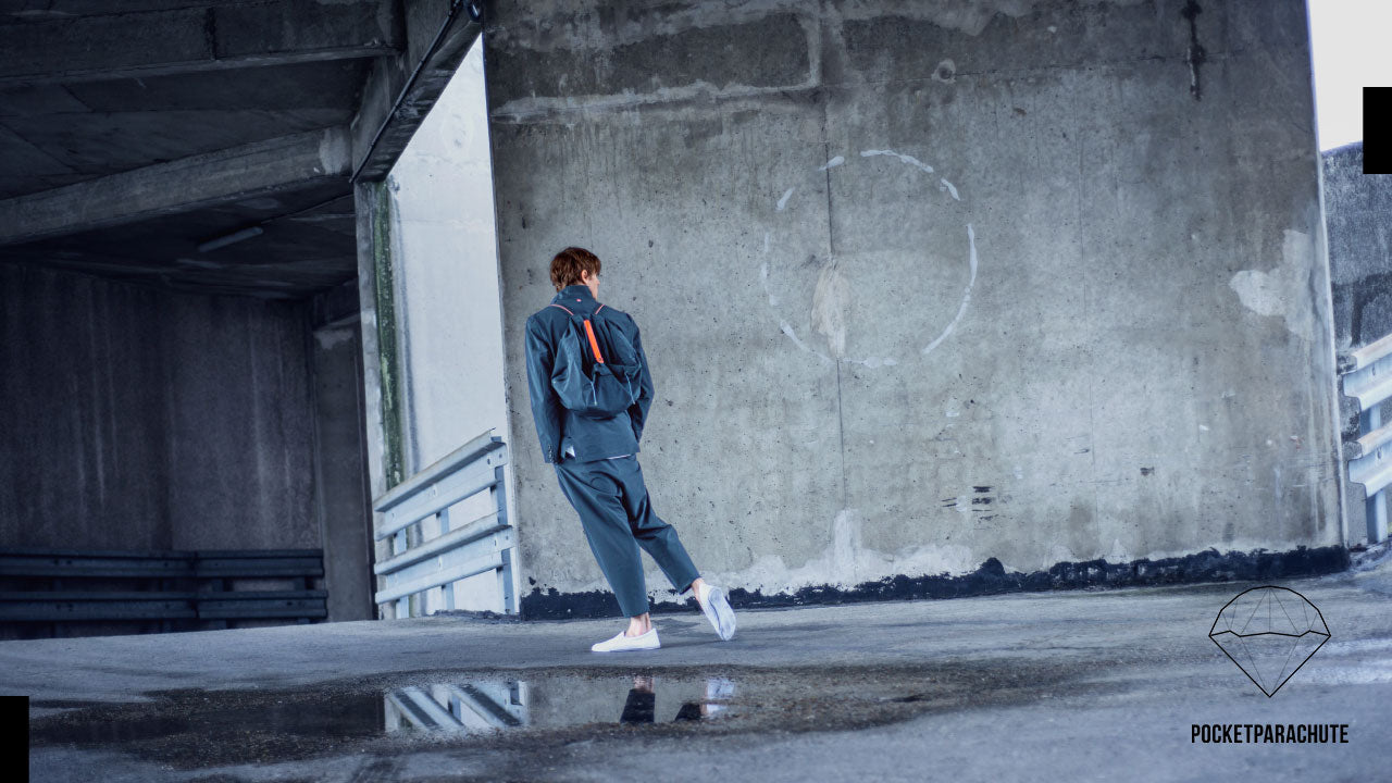 A person walking on a wet concrete surface in an urban setting, wearing a dark jacket, pants, and white sneakers. They are carrying a small backpack. The background features a large concrete structure with the logo POCKETPARACHUTE.