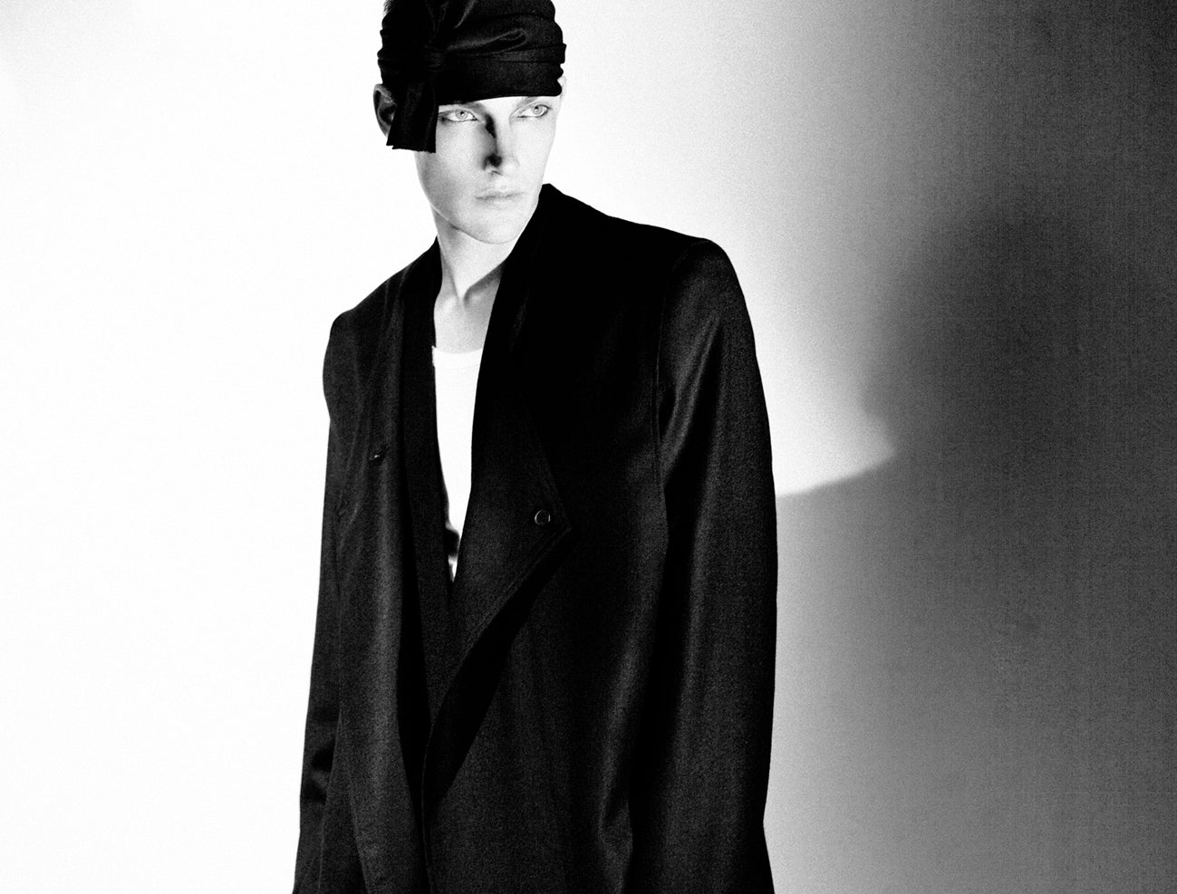 A person wearing a large black blazer over a white shirt stands against a bright background. They have a dark cloth tied around their head, covering one eye. The image has a high contrast, black and white effect.